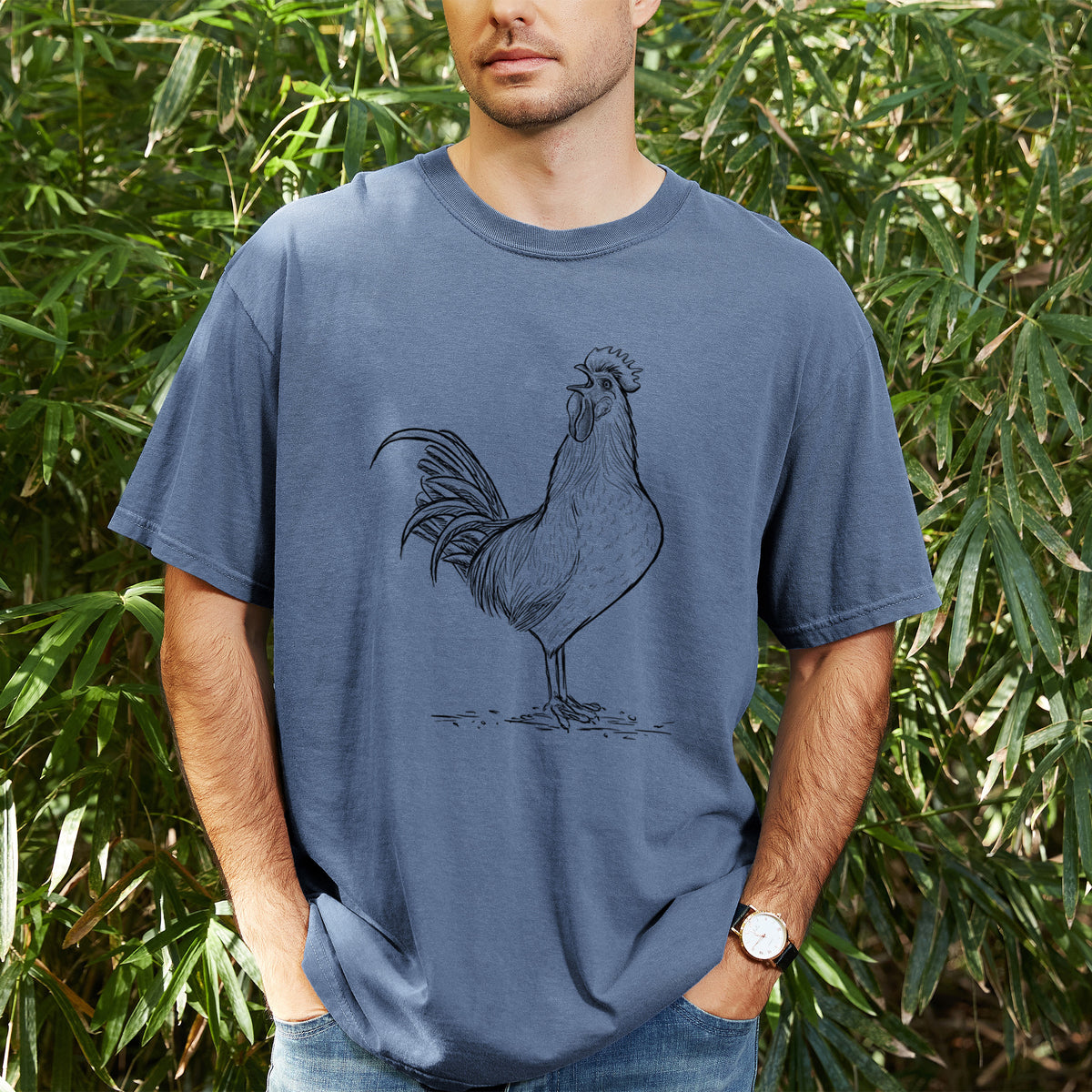 Man wearing a Comfort Colors t-shirt printed with a detailed black ink illustration of a rooster crowing, outdoors.