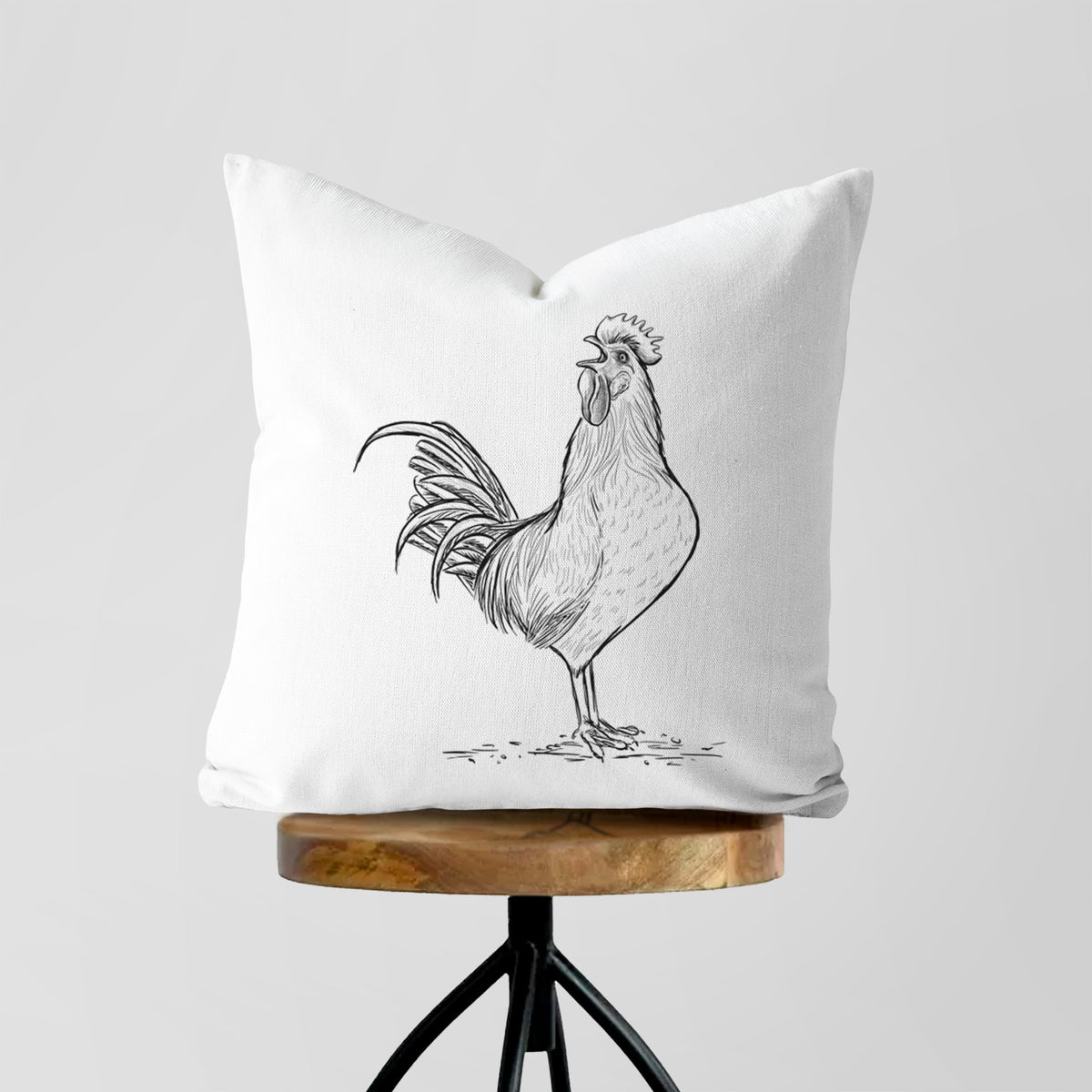 White pillow cover printed with a detailed black ink illustration of a rooster crowing, on a wooden stool.