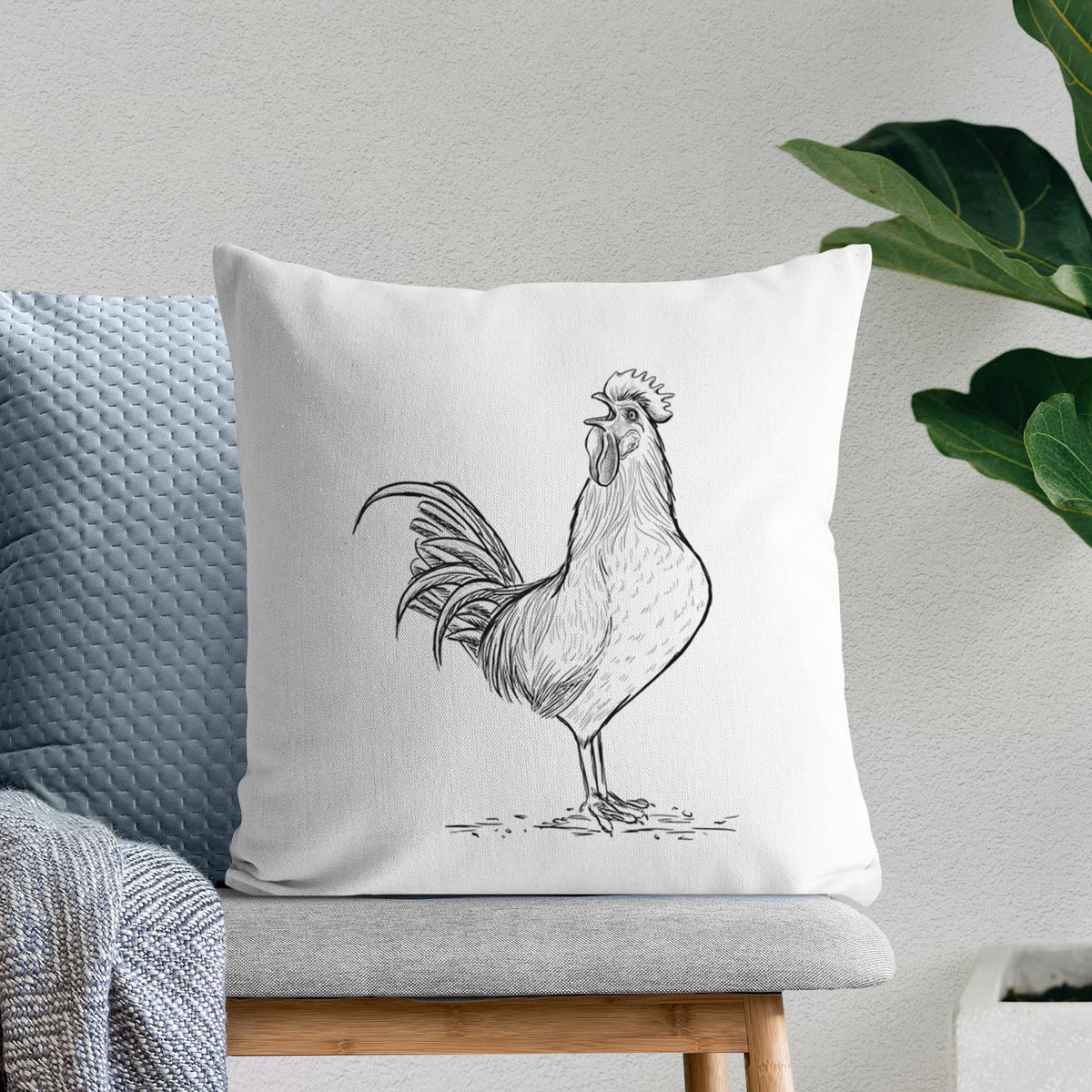 White pillow cover printed with a detailed black ink illustration of a rooster crowing, on a bench beside plants.