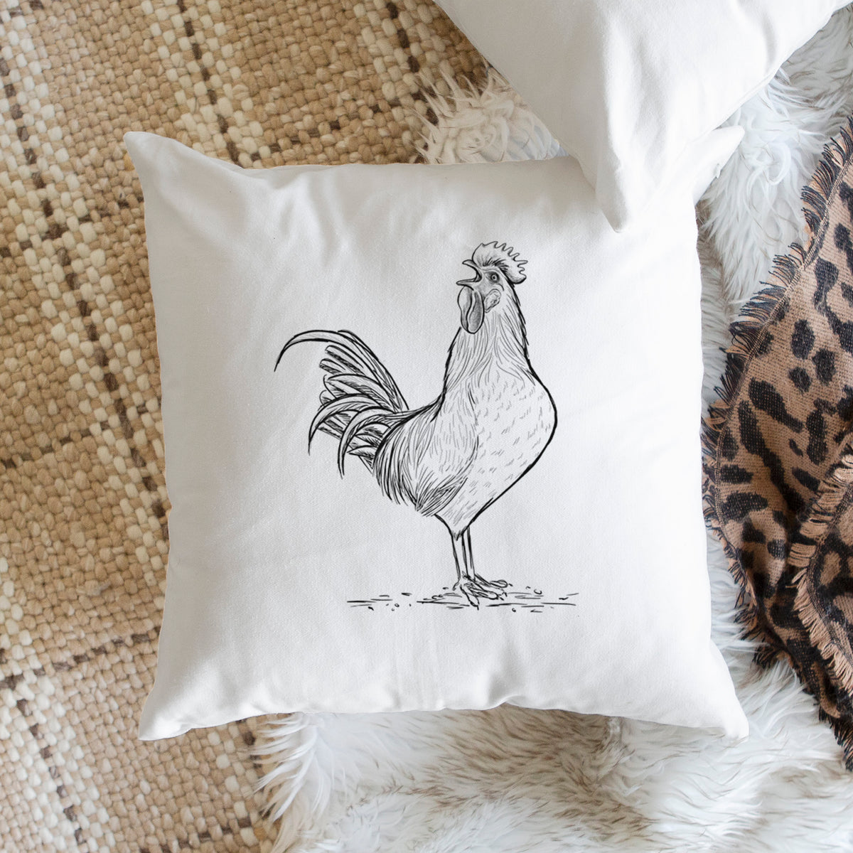 White pillow cover printed with a detailed black ink illustration of a rooster crowing, on a woven chair.