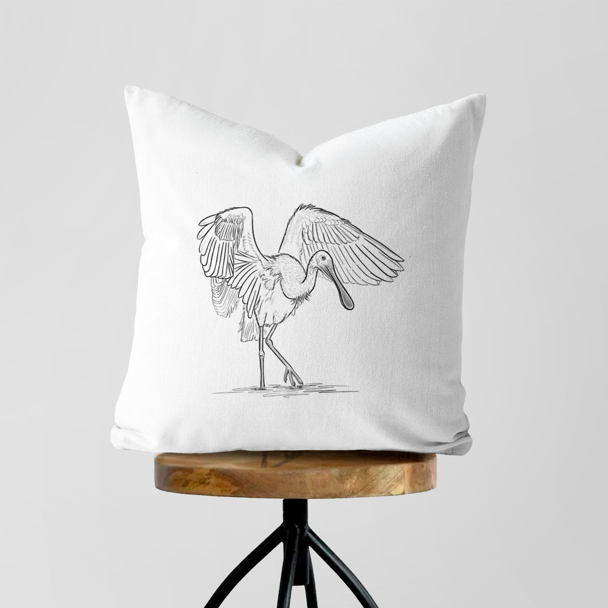 White pillow cover printed with a black ink illustration of a roseate spoonbill standing with its wings spread, on a wooden stool.