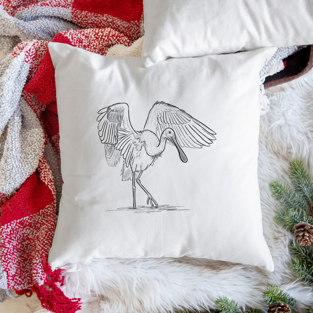 White pillow cover printed with a black ink illustration of a roseate spoonbill standing with its wings spread, on a woven chair.