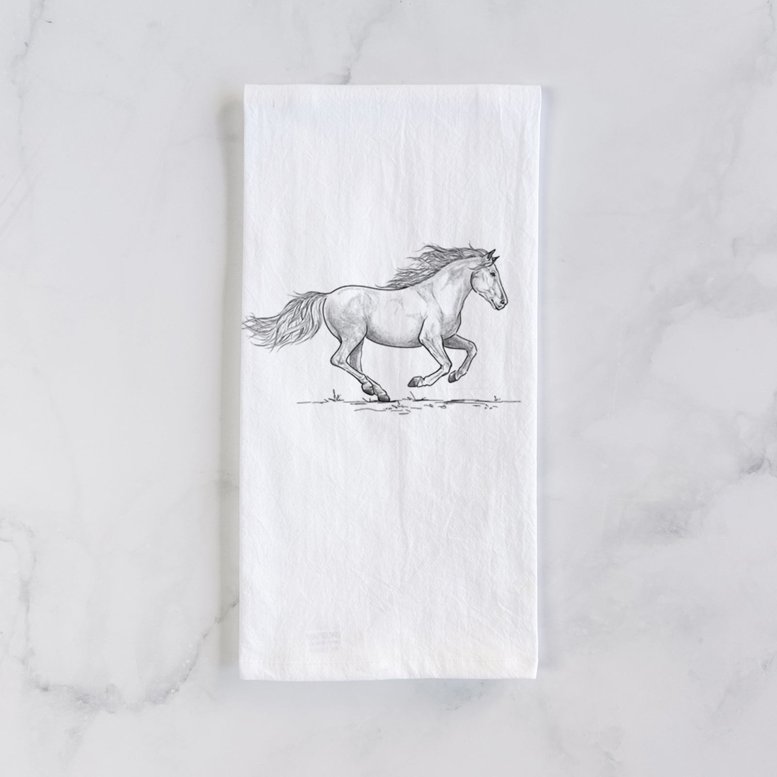 White cotton tea towel printed with a black ink illustration of a mustang horse running, on a marble counter beside a round wooden cutting board and greenery.