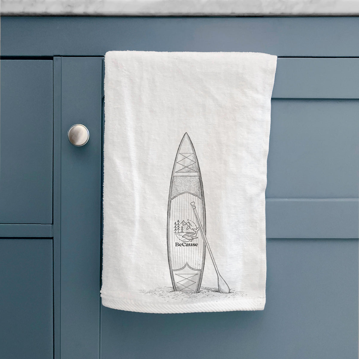 White hand towel printed with a black ink illustration of a stand-up paddleboard with a paddle, hanging on a cabinet door.
