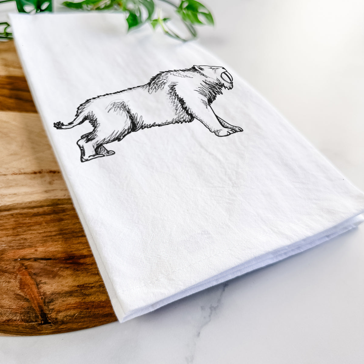 Close-up of a folded white tea towel on a wooden board, showing a black ink illustration of a saber-toothed tiger walking.