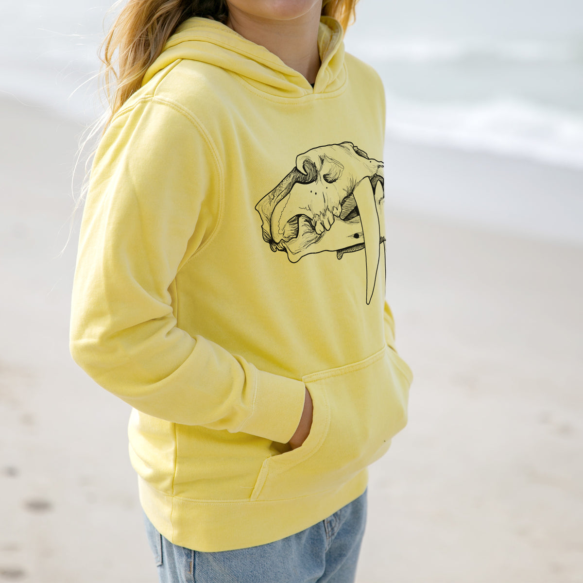 Girl wearing a youth pigment dyed hoodie printed with a detailed black ink illustration of a saber-toothed tiger skull.
