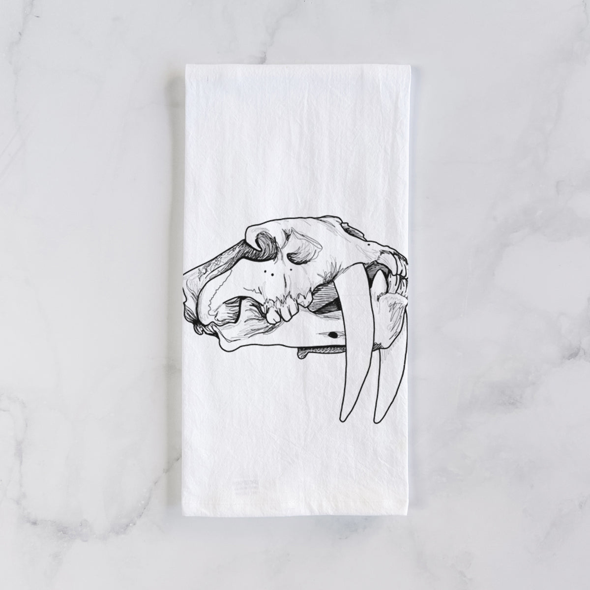 White cotton tea towel laid flat on marble, printed with a detailed black ink illustration of a saber-toothed tiger skull.