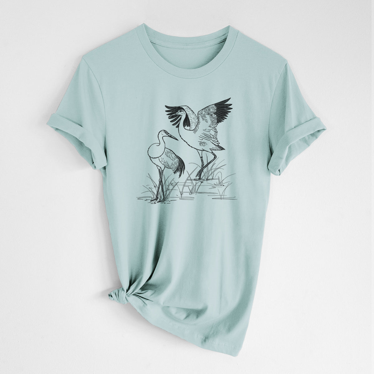 Dusty blue lightweight cotton t-shirt laid flat with the sleeves rolled, printed with a black ink illustration of two sandhill cranes in marsh grass, one with its wings raised.