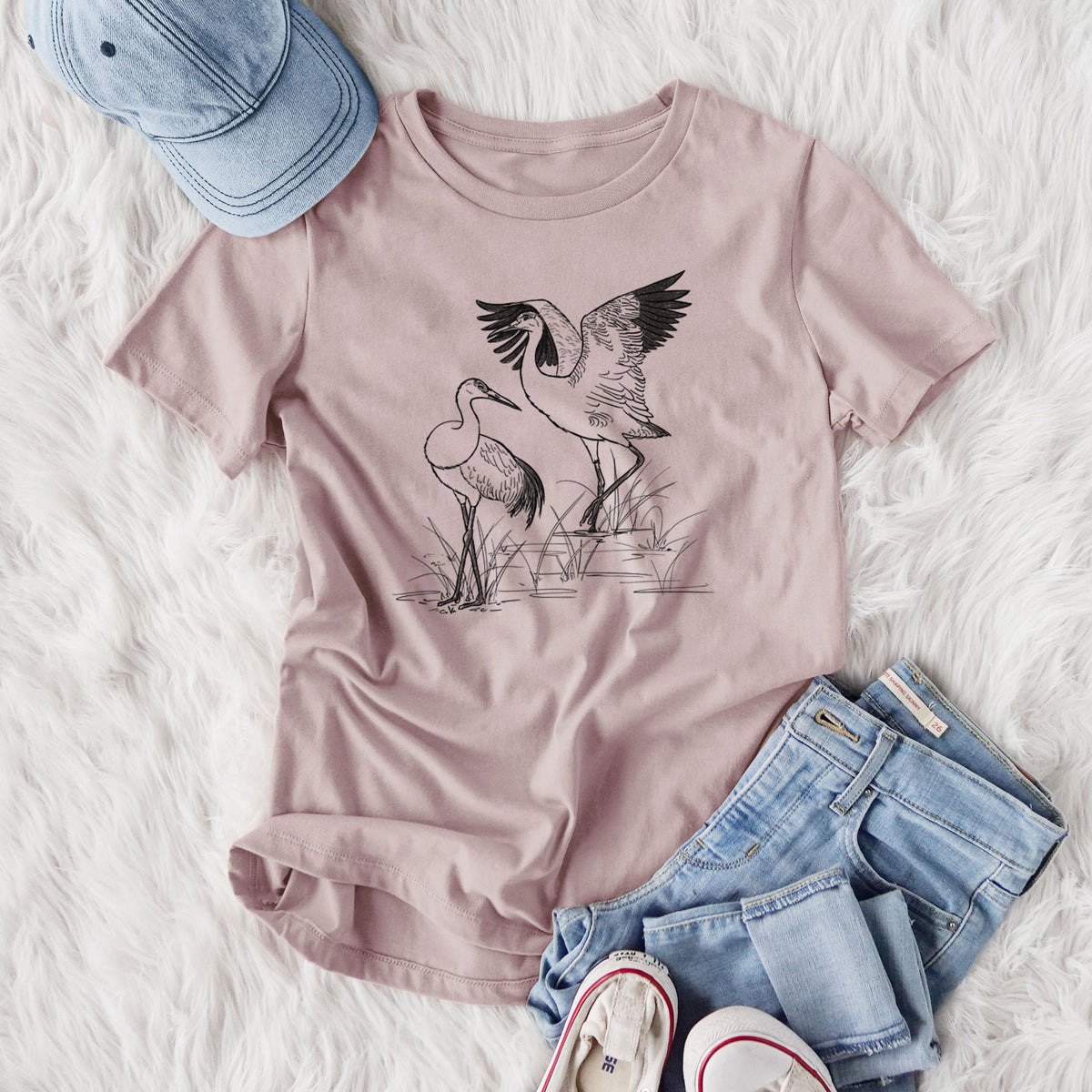 Women's relaxed fit cotton t-shirt printed with a black ink illustration of two sandhill cranes in marsh grass, one with its wings raised, styled flat on a white faux fur rug with jeans.
