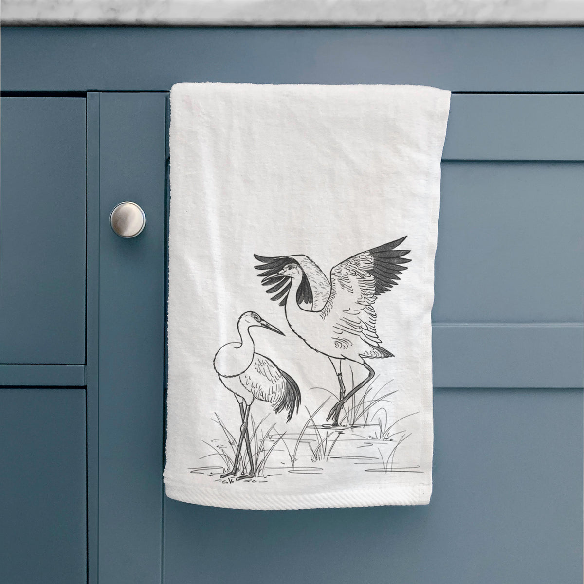 White hand towel printed with a black ink illustration of two sandhill cranes in marsh grass, one with its wings raised, hanging on a cabinet door.