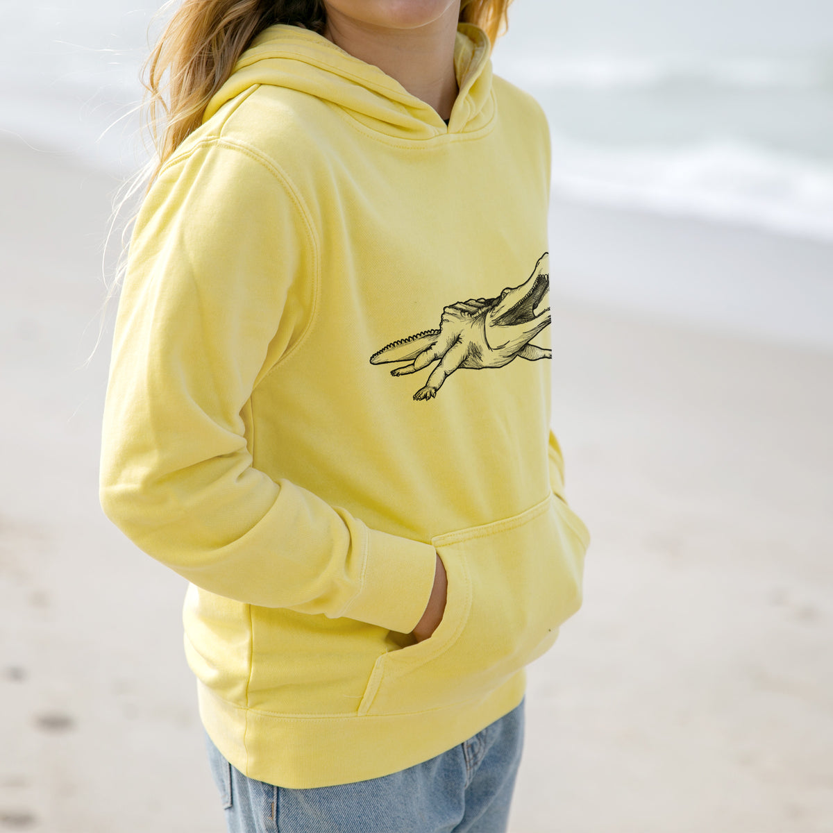 Girl wearing a youth pigment dyed hoodie printed with a black ink illustration of a Sarcosuchus, a giant prehistoric crocodile.