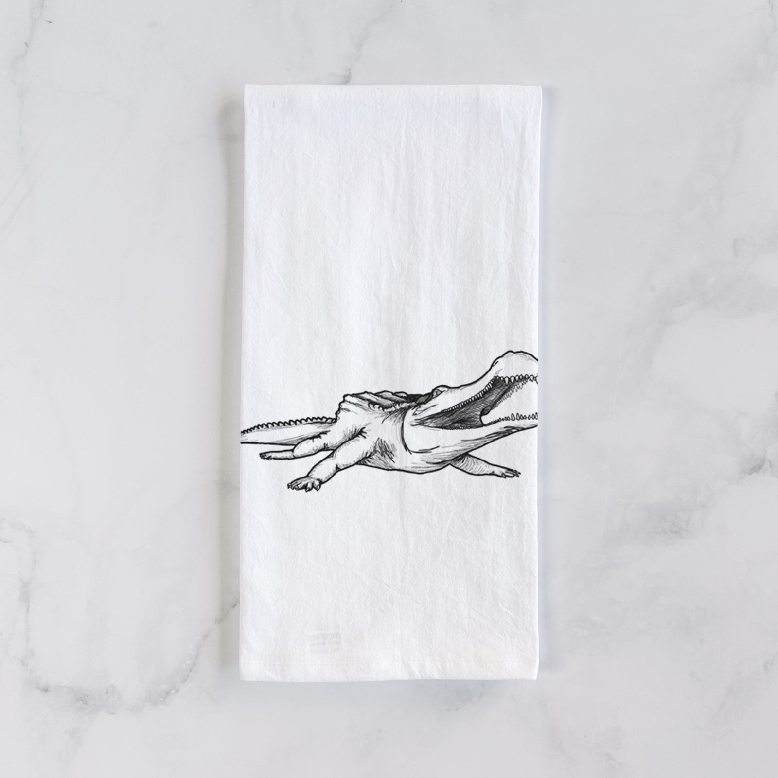 White cotton tea towel printed with a black ink illustration of a Sarcosuchus, a giant prehistoric crocodile, on a marble counter beside a round wooden cutting board and greenery.