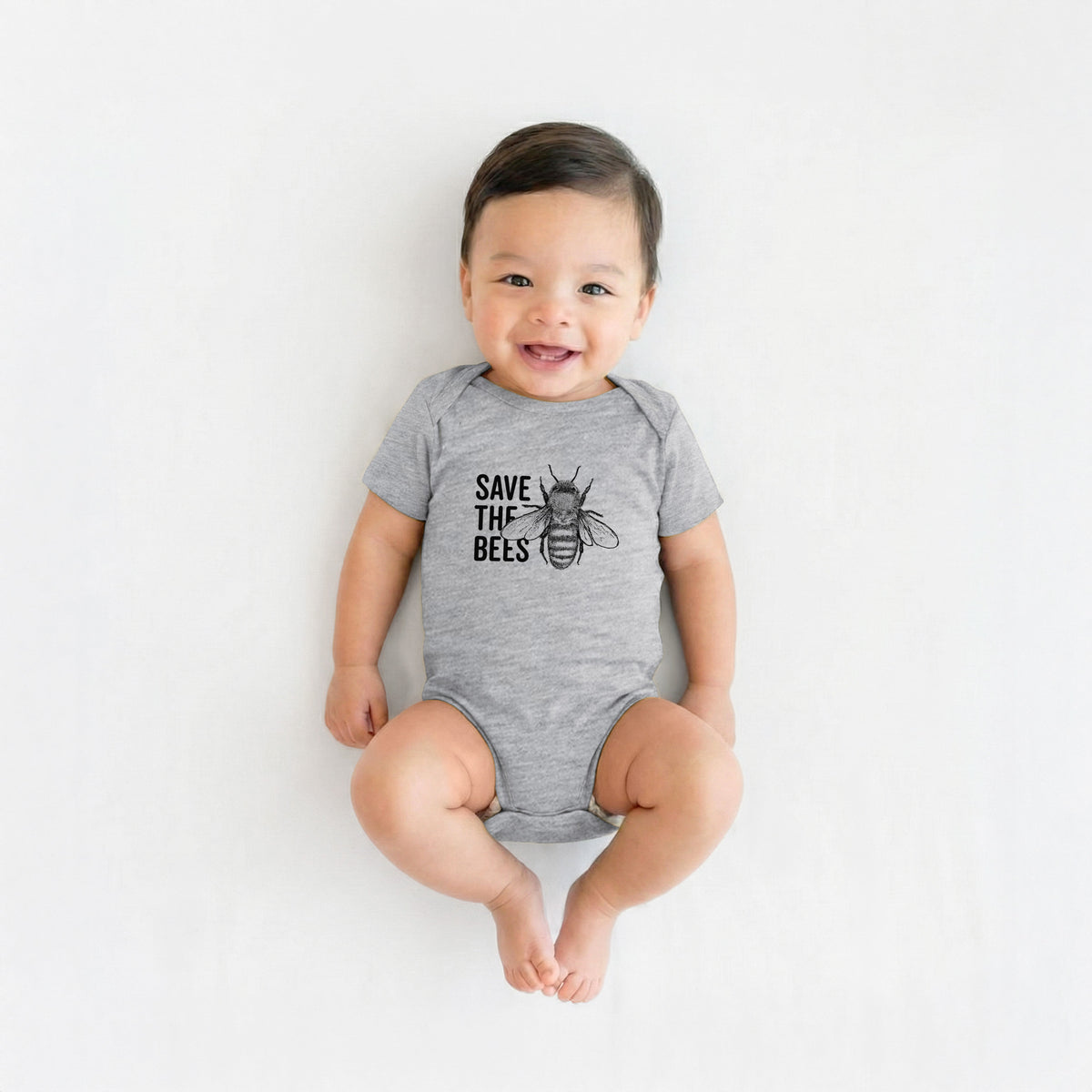 Smiling baby boy wearing a grey baby bodysuit printed with bold black words reading Save the Bees beside a detailed honey bee illustration.