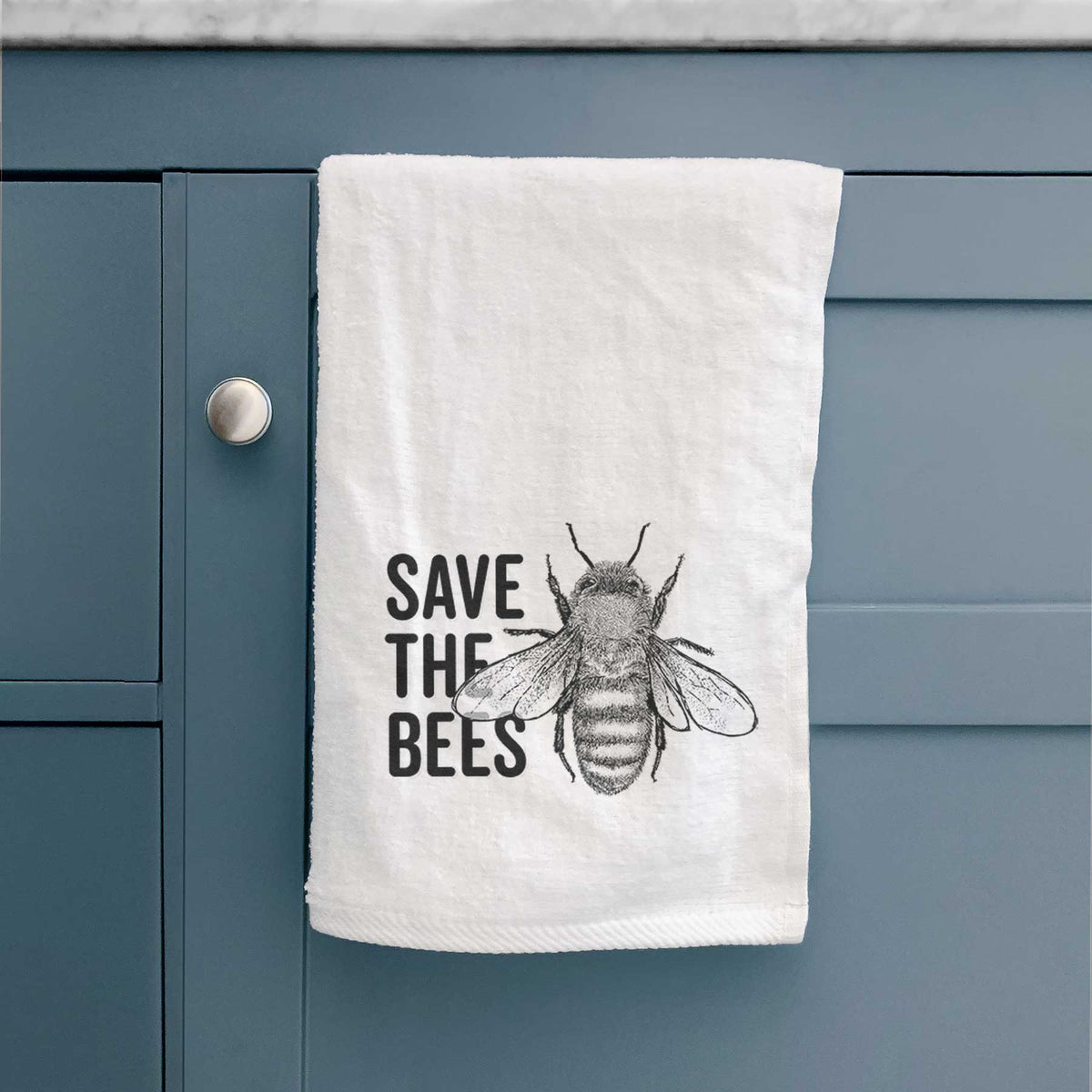 White hand towel printed with bold black words reading Save the Bees beside a detailed honey bee illustration, hanging on a cabinet door.