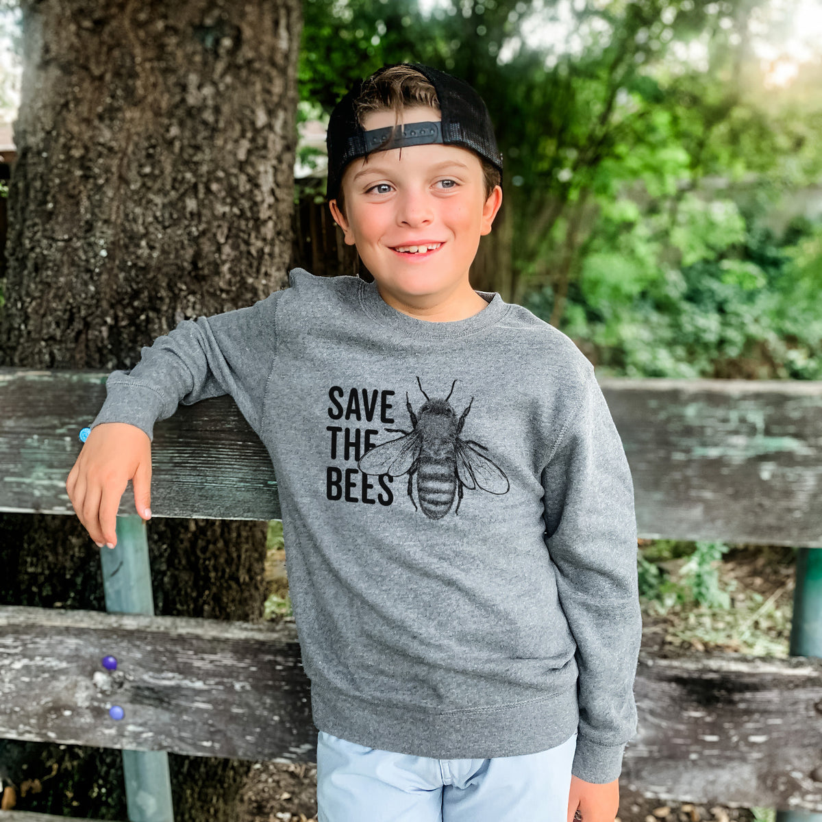 Boy wearing a youth crewneck sweatshirt printed with bold black words reading Save the Bees beside a detailed honey bee illustration, outdoors.