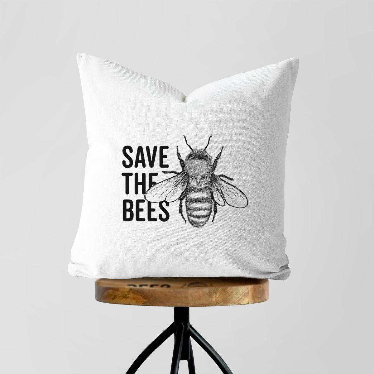 White pillow cover printed with bold black words reading Save the Bees beside a detailed honey bee illustration, on a wooden stool.