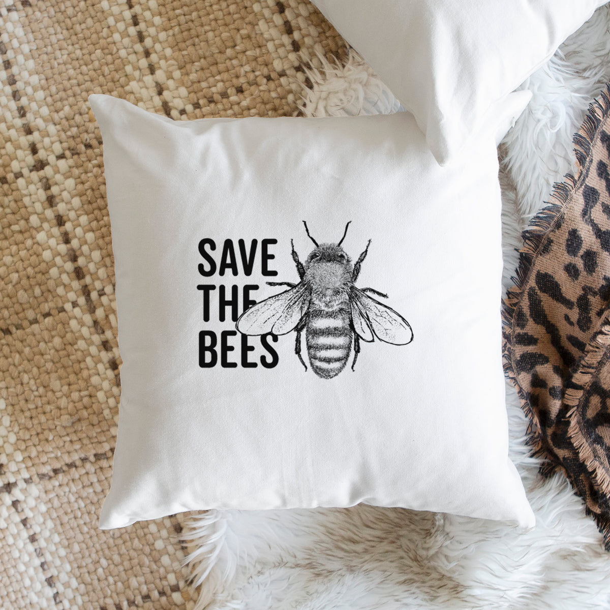White pillow cover printed with bold black words reading Save the Bees beside a detailed honey bee illustration, on a woven chair.