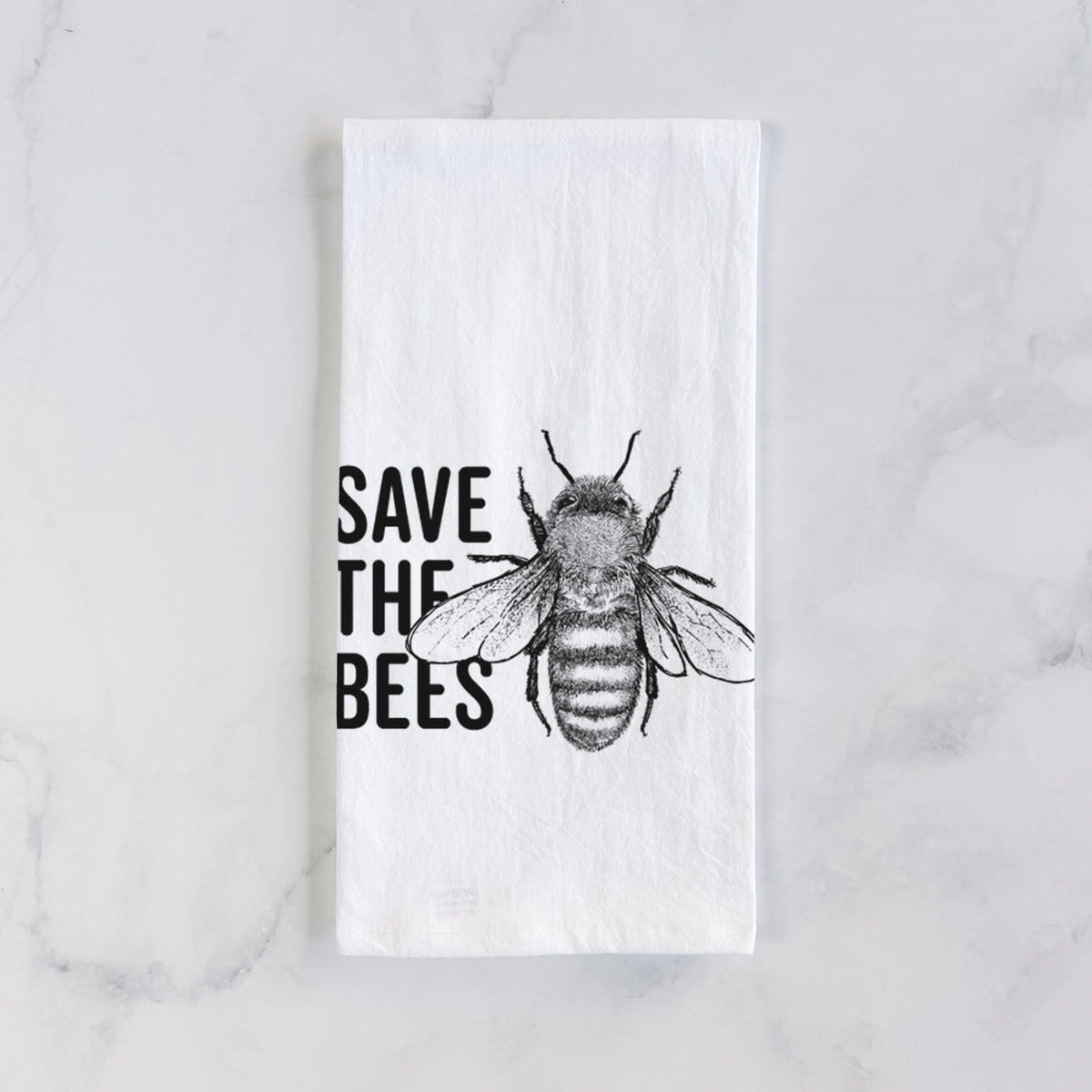 White cotton tea towel laid flat on marble, printed with bold black words reading Save the Bees beside a detailed honey bee illustration.