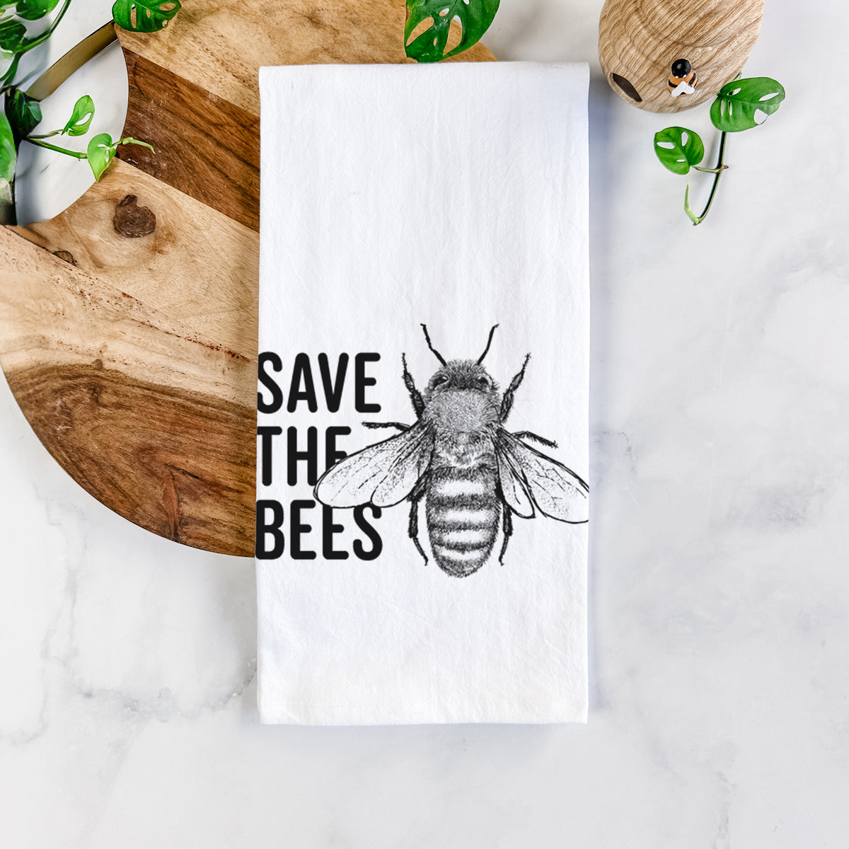 White cotton tea towel printed with bold black words reading Save the Bees beside a detailed honey bee illustration, on a marble counter beside a round wooden cutting board and greenery.