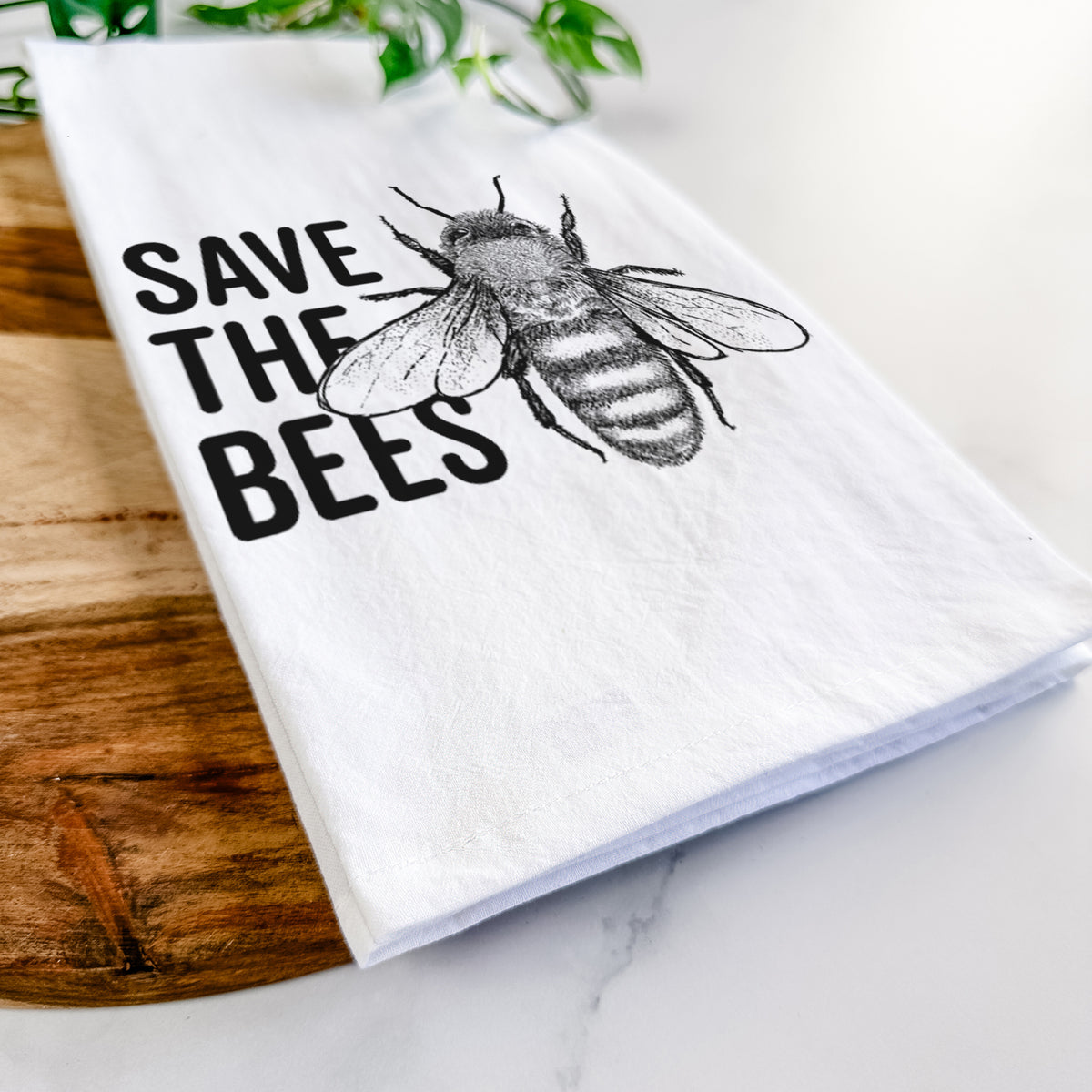 Close-up of a folded white tea towel on a wooden board, showing bold black words reading Save the Bees beside a detailed honey bee illustration.