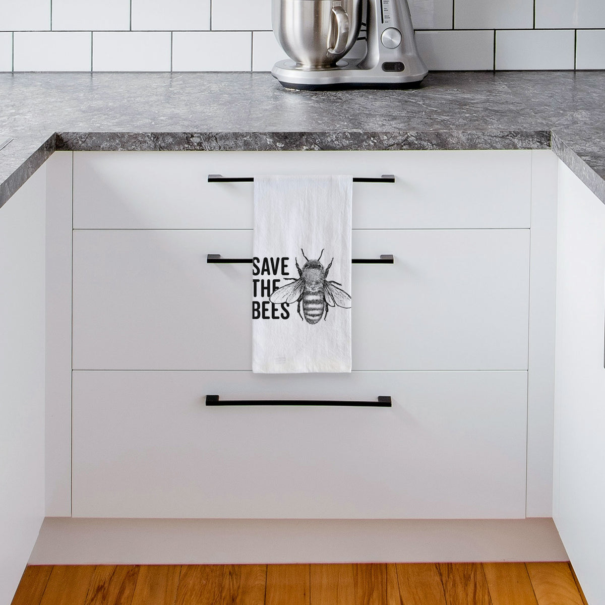 White tea towel printed with bold black words reading Save the Bees beside a detailed honey bee illustration, hanging from a drawer handle in a bright white kitchen.