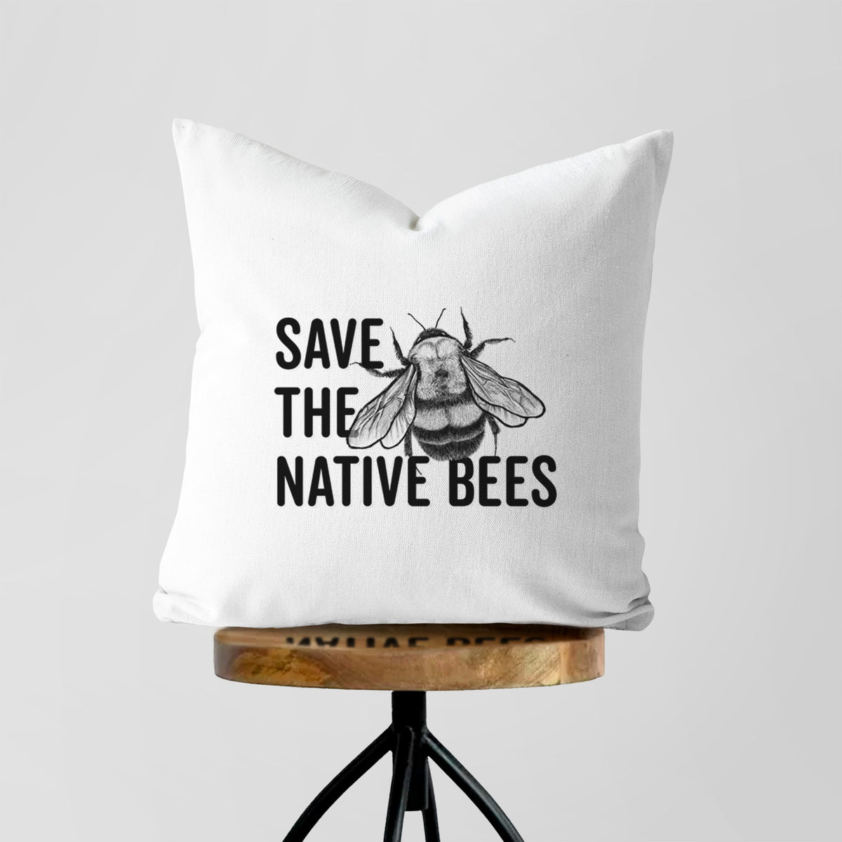 White pillow cover printed with bold black words reading Save the Native Bees beside a detailed bee illustration, on a wooden stool.