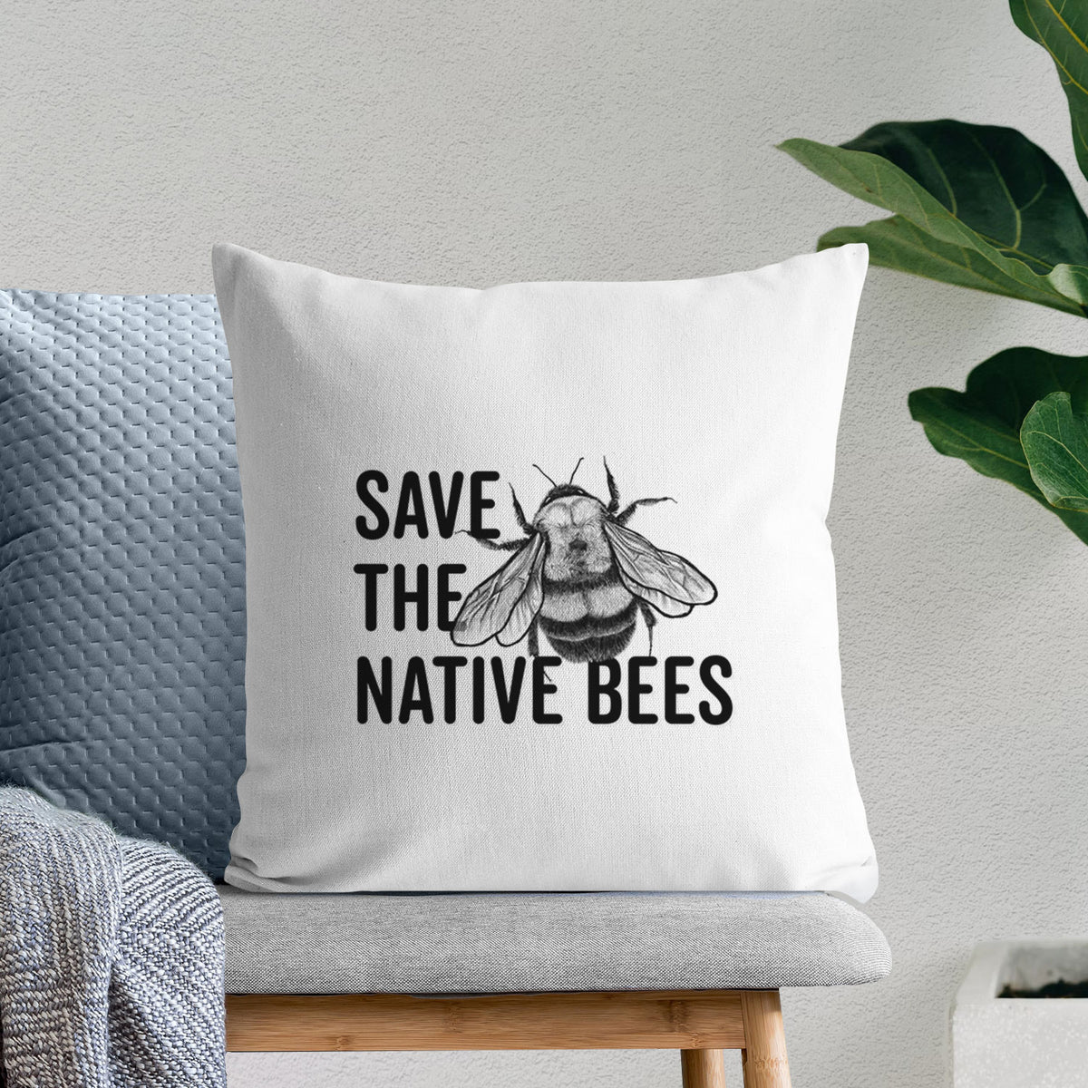 White pillow cover printed with bold black words reading Save the Native Bees beside a detailed bee illustration, on a bench beside plants.