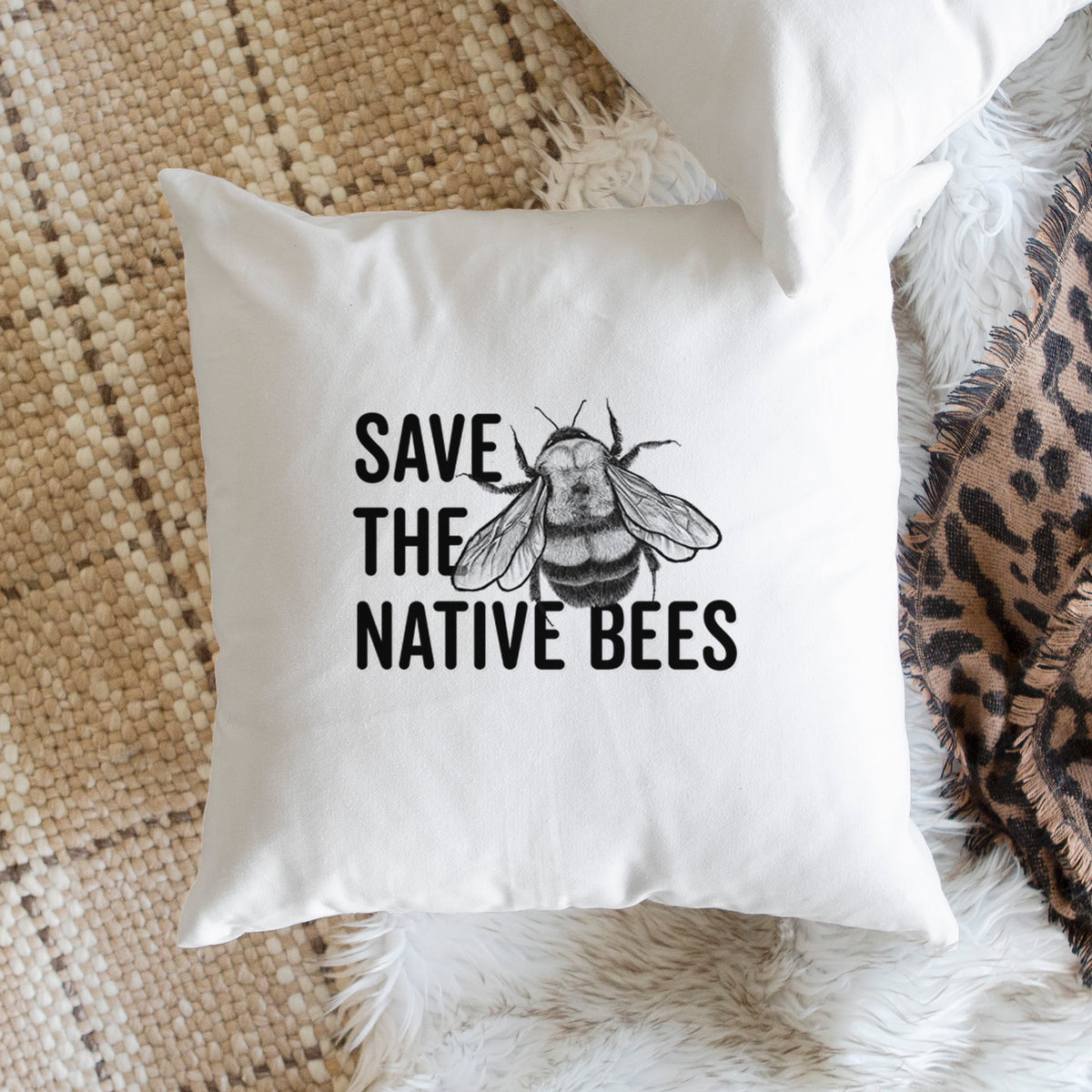 White pillow cover printed with bold black words reading Save the Native Bees beside a detailed bee illustration, on a woven chair.