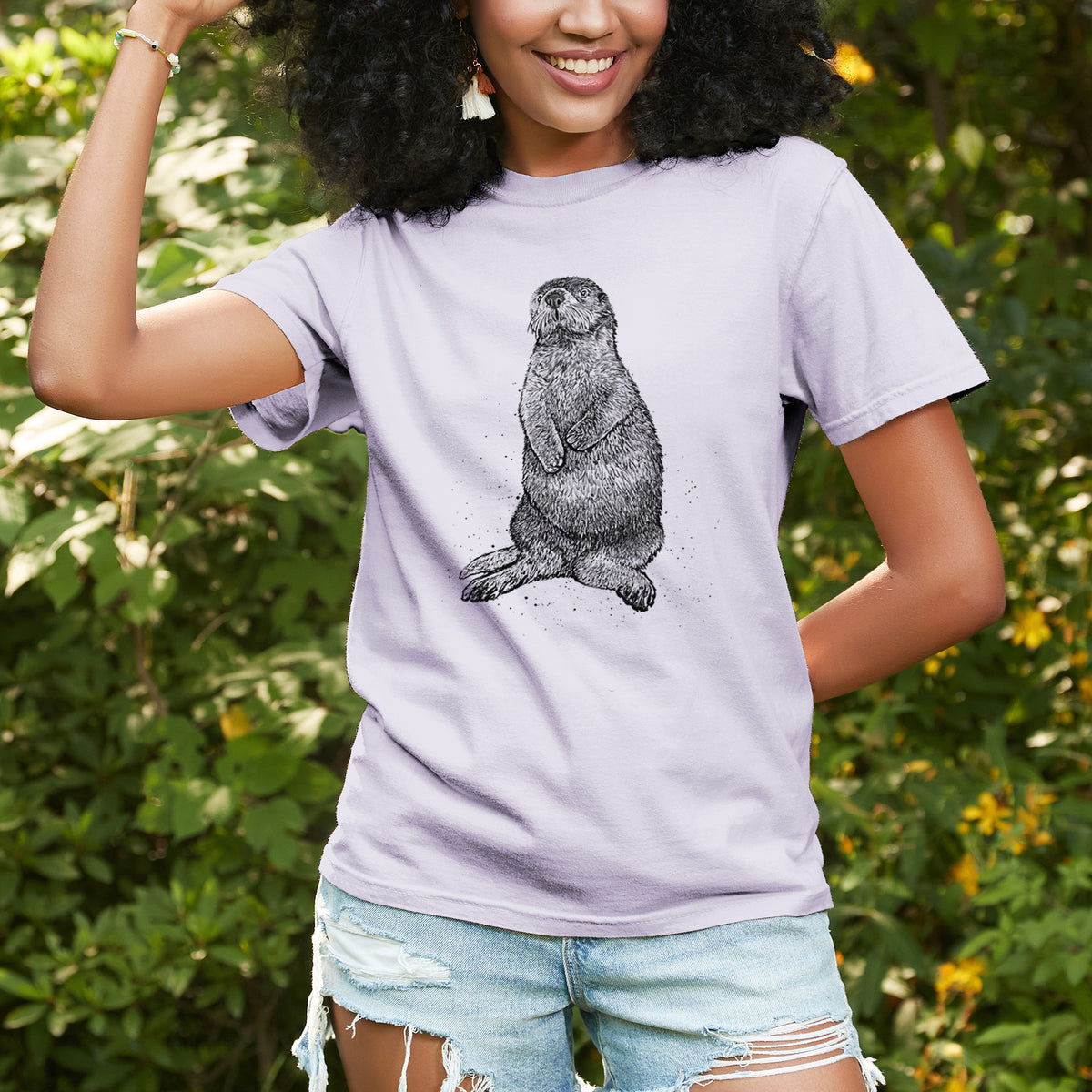 Woman wearing a Comfort Colors t-shirt printed with a detailed black ink illustration of a sea otter sitting upright, outdoors.