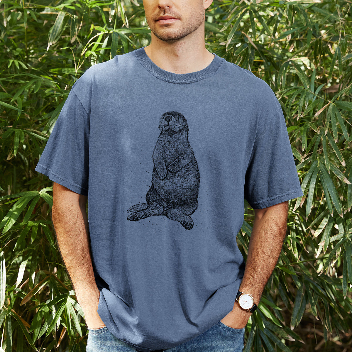 Man wearing a Comfort Colors t-shirt printed with a detailed black ink illustration of a sea otter sitting upright, outdoors.