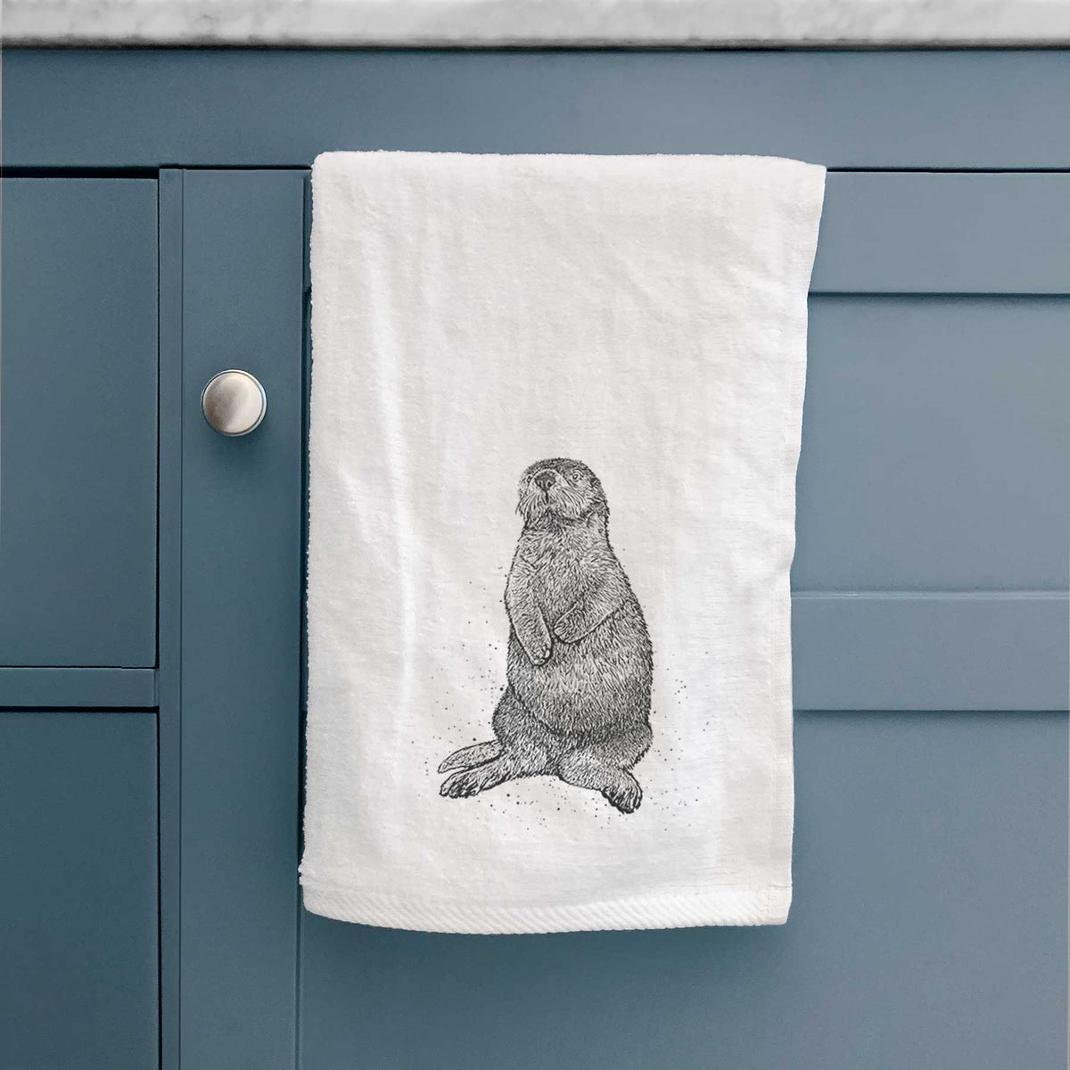 White hand towel printed with a detailed black ink illustration of a sea otter sitting upright, hanging on a cabinet door.