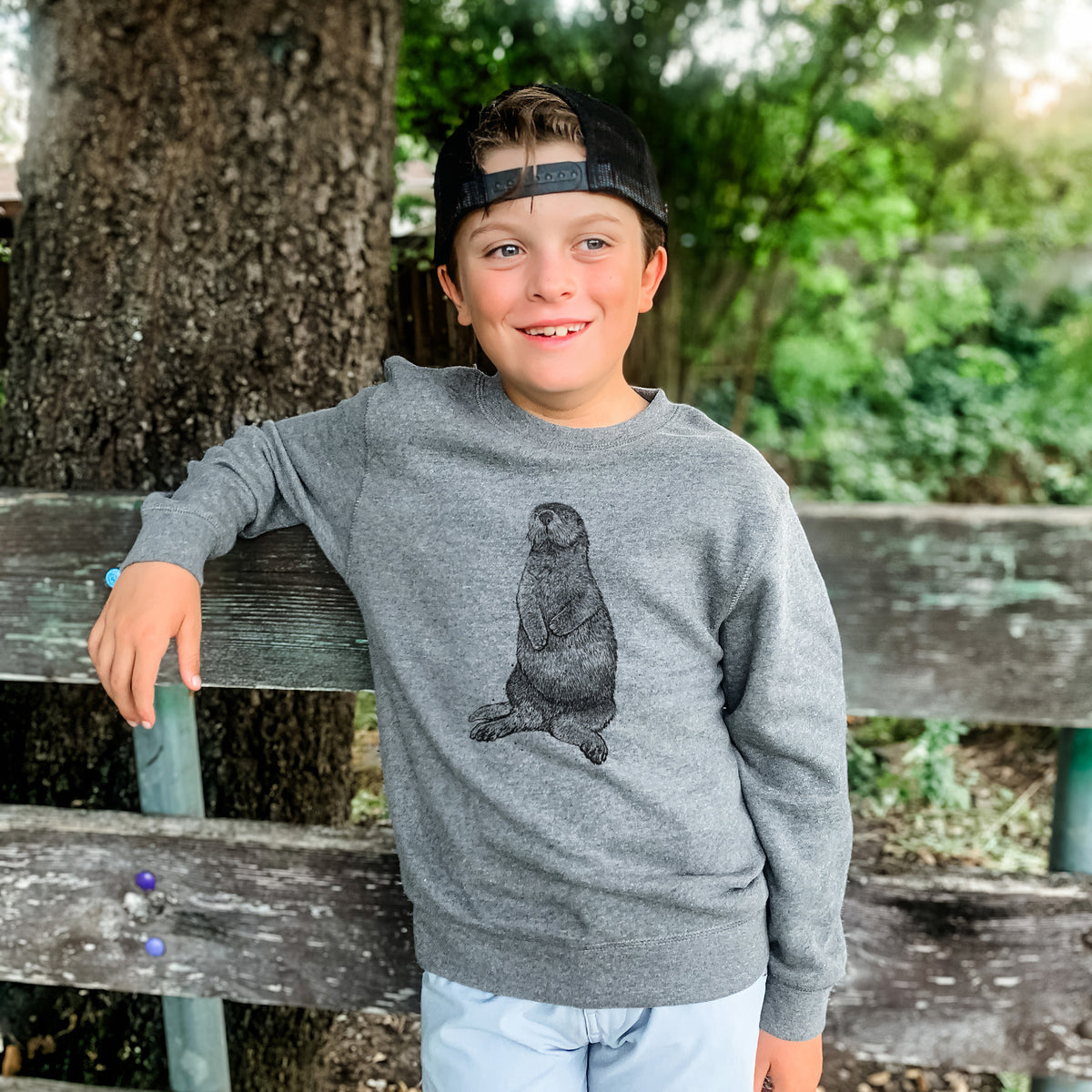 Boy wearing a youth crewneck sweatshirt printed with a detailed black ink illustration of a sea otter sitting upright, outdoors.
