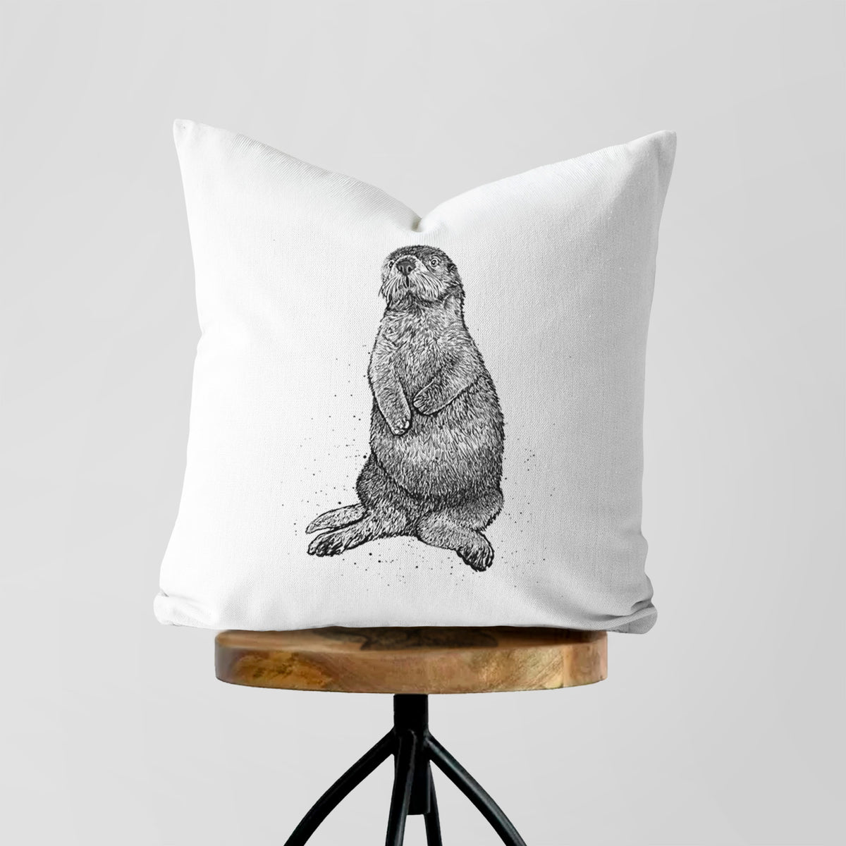 White pillow cover printed with a detailed black ink illustration of a sea otter sitting upright, on a wooden stool.