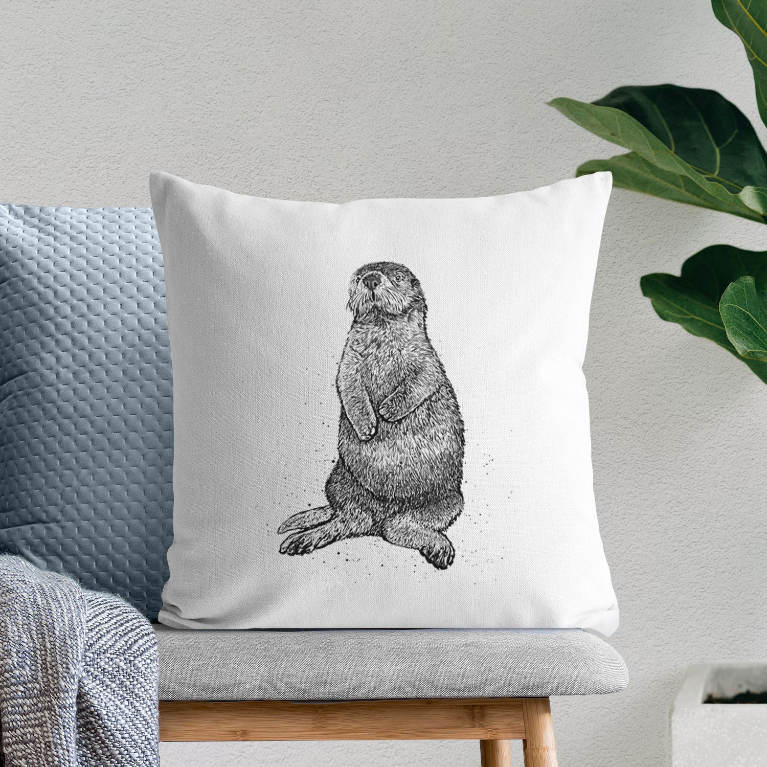 White pillow cover printed with a detailed black ink illustration of a sea otter sitting upright, on a bench beside plants.