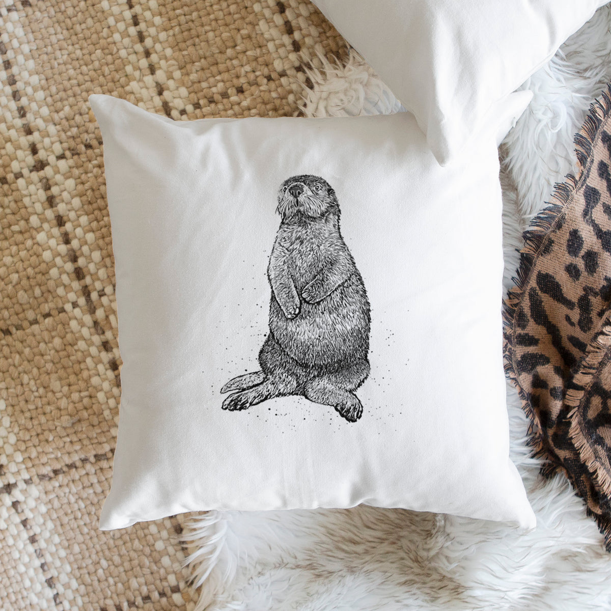 White pillow cover printed with a detailed black ink illustration of a sea otter sitting upright, on a woven chair.