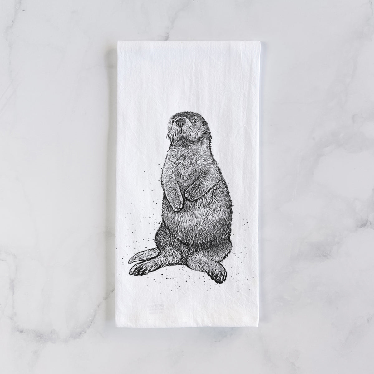 White cotton tea towel laid flat on marble, printed with a detailed black ink illustration of a sea otter sitting upright.