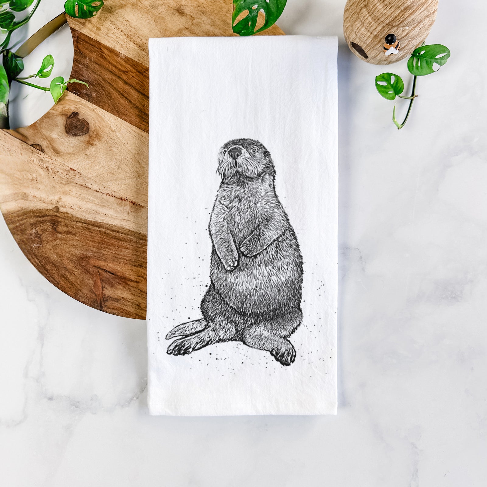 White cotton tea towel printed with a detailed black ink illustration of a sea otter sitting upright, on a marble counter beside a round wooden cutting board and greenery.