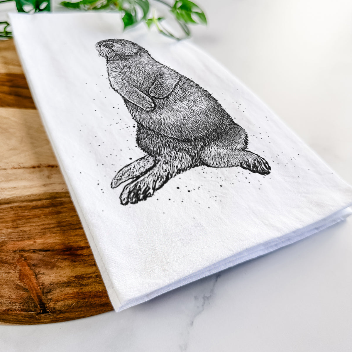 Close-up of a folded white tea towel on a wooden board, showing a detailed black ink illustration of a sea otter sitting upright.