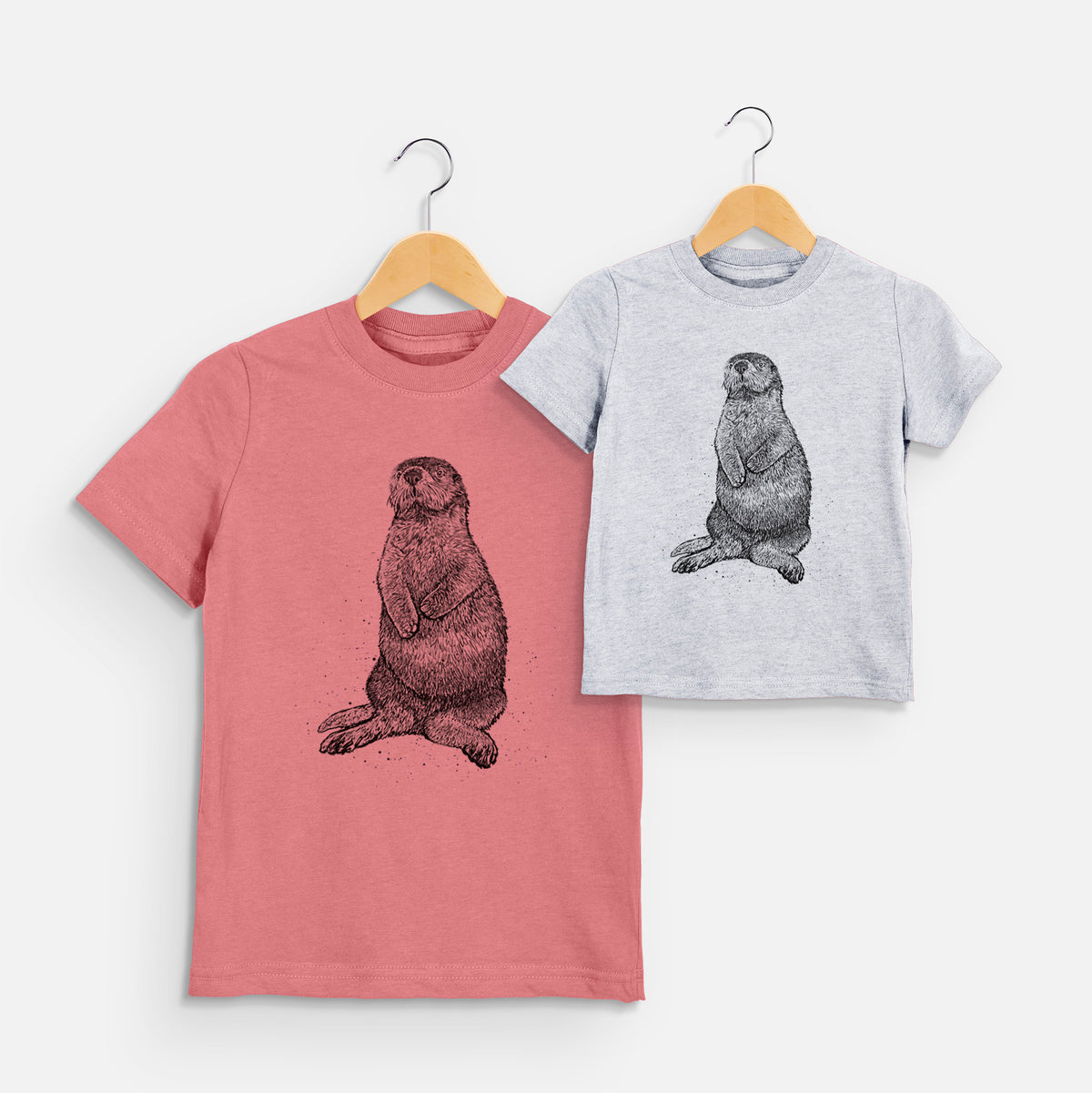 Two youth t-shirts in different colors on hangers, printed with a detailed black ink illustration of a sea otter sitting upright.