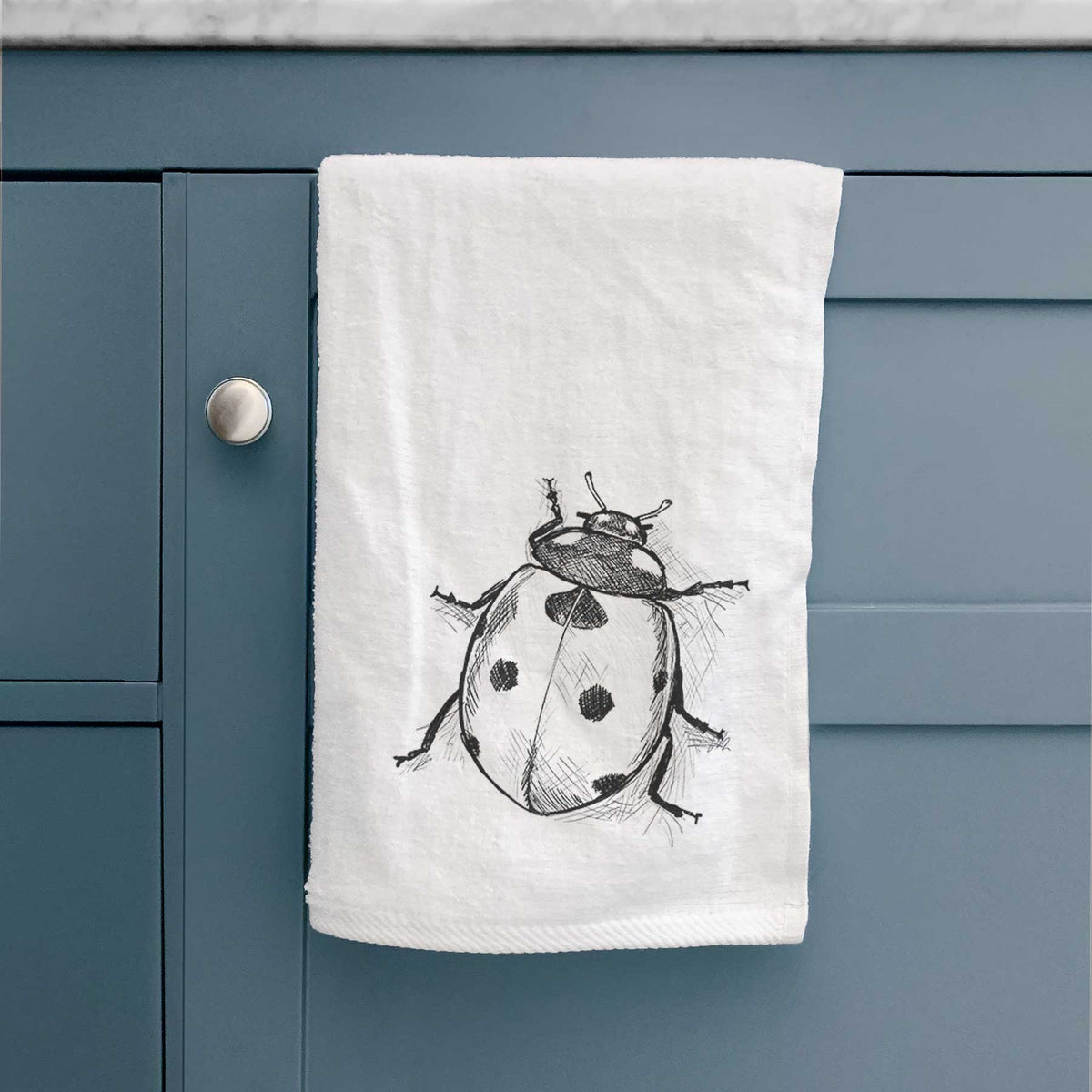White hand towel printed with a detailed black ink illustration of a seven-spot ladybird beetle, hanging on a cabinet door.