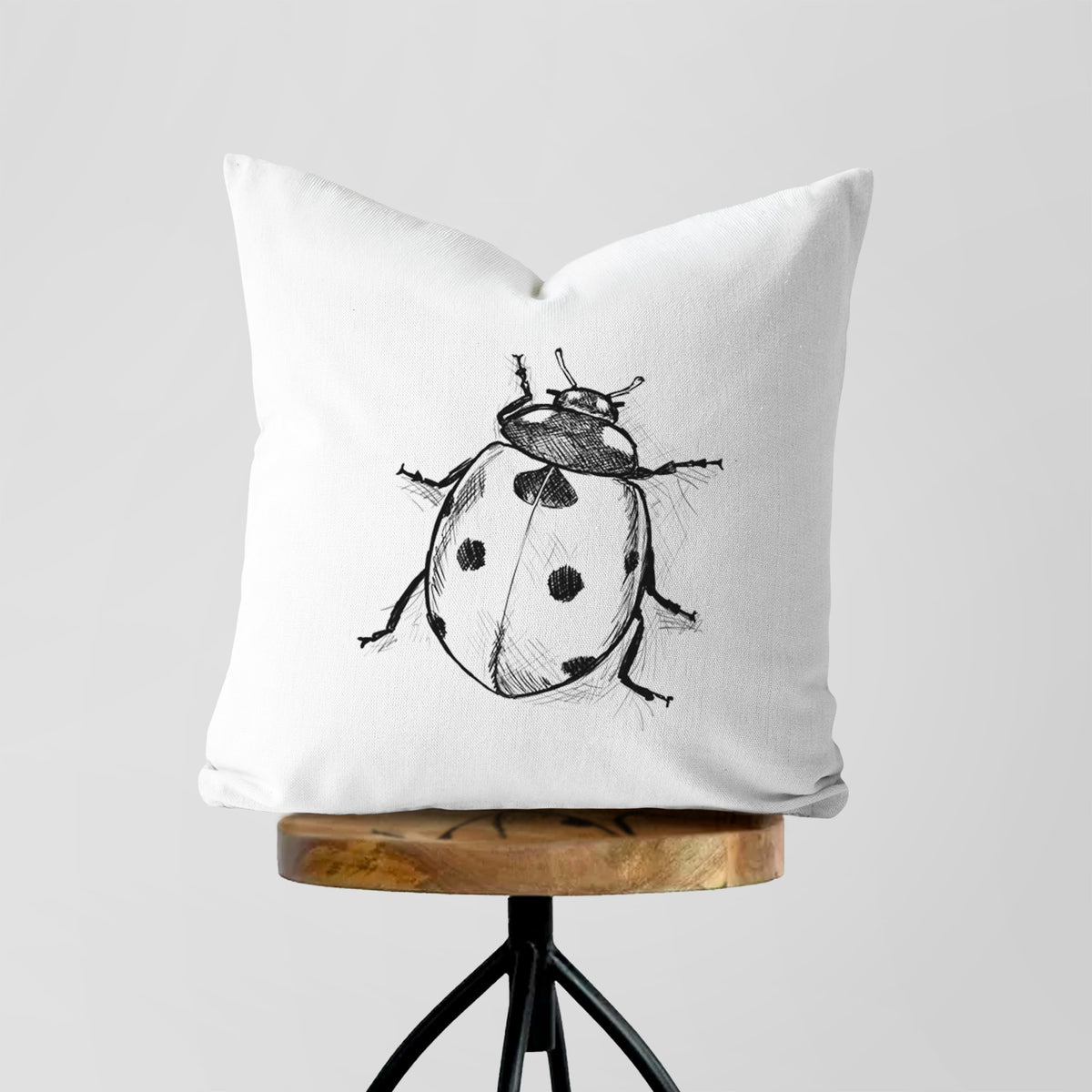White pillow cover printed with a detailed black ink illustration of a seven-spot ladybird beetle, on a wooden stool.