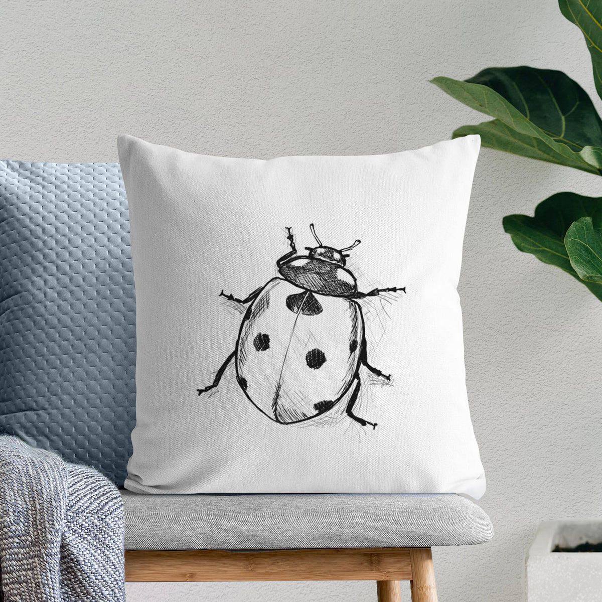 White pillow cover printed with a detailed black ink illustration of a seven-spot ladybird beetle, on a bench beside plants.