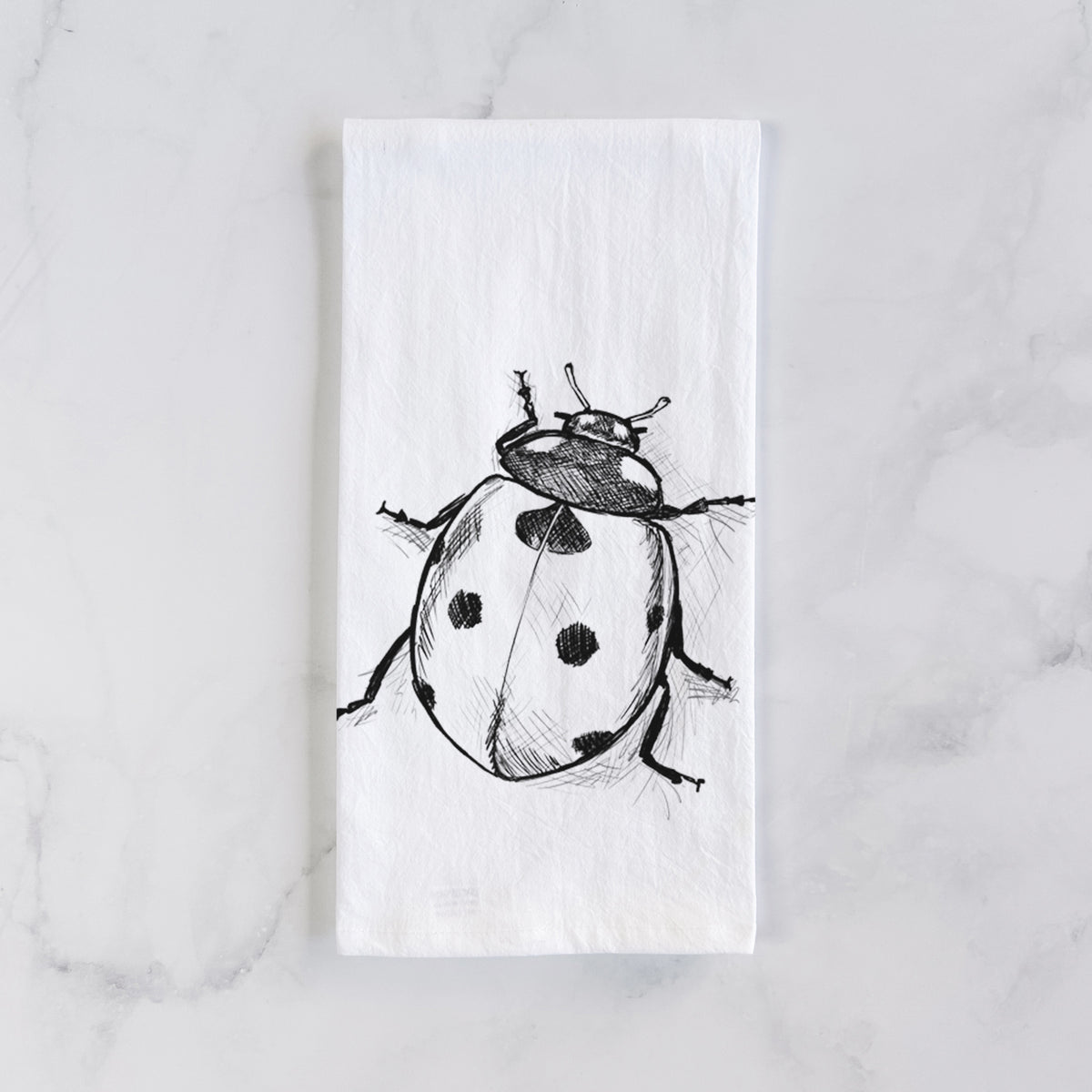 White cotton tea towel laid flat on marble, printed with a detailed black ink illustration of a seven-spot ladybird beetle.