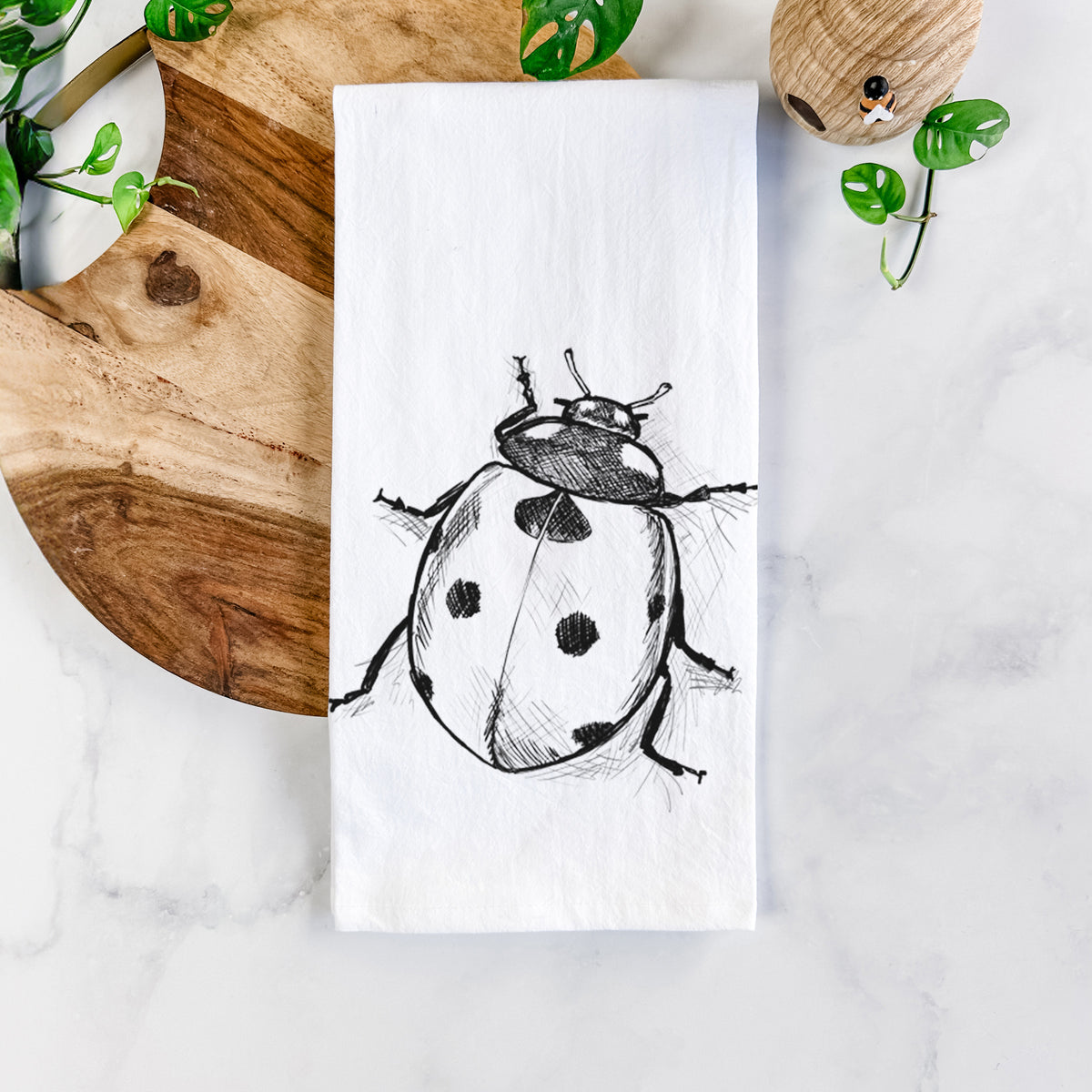 White cotton tea towel printed with a detailed black ink illustration of a seven-spot ladybird beetle, on a marble counter beside a round wooden cutting board and greenery.