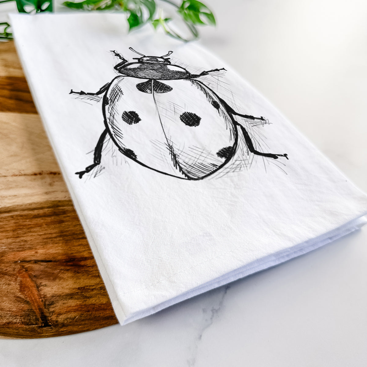 Close-up of a folded white tea towel on a wooden board, showing a detailed black ink illustration of a seven-spot ladybird beetle.