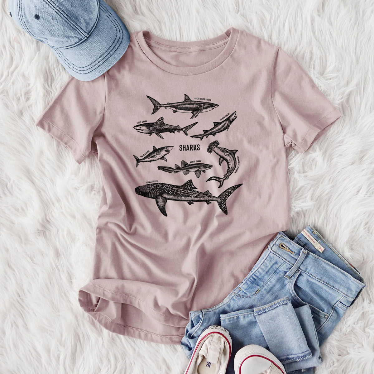 Women's relaxed fit cotton t-shirt printed with a chart of black ink shark illustrations, including a whale shark, hammerhead and great white, styled flat on a white faux fur rug with jeans.