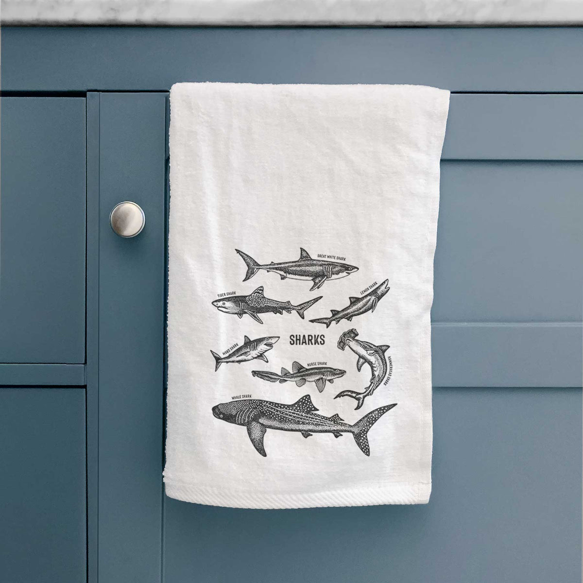 White hand towel printed with a chart of black ink shark illustrations, including a whale shark, hammerhead and great white, hanging on a cabinet door.