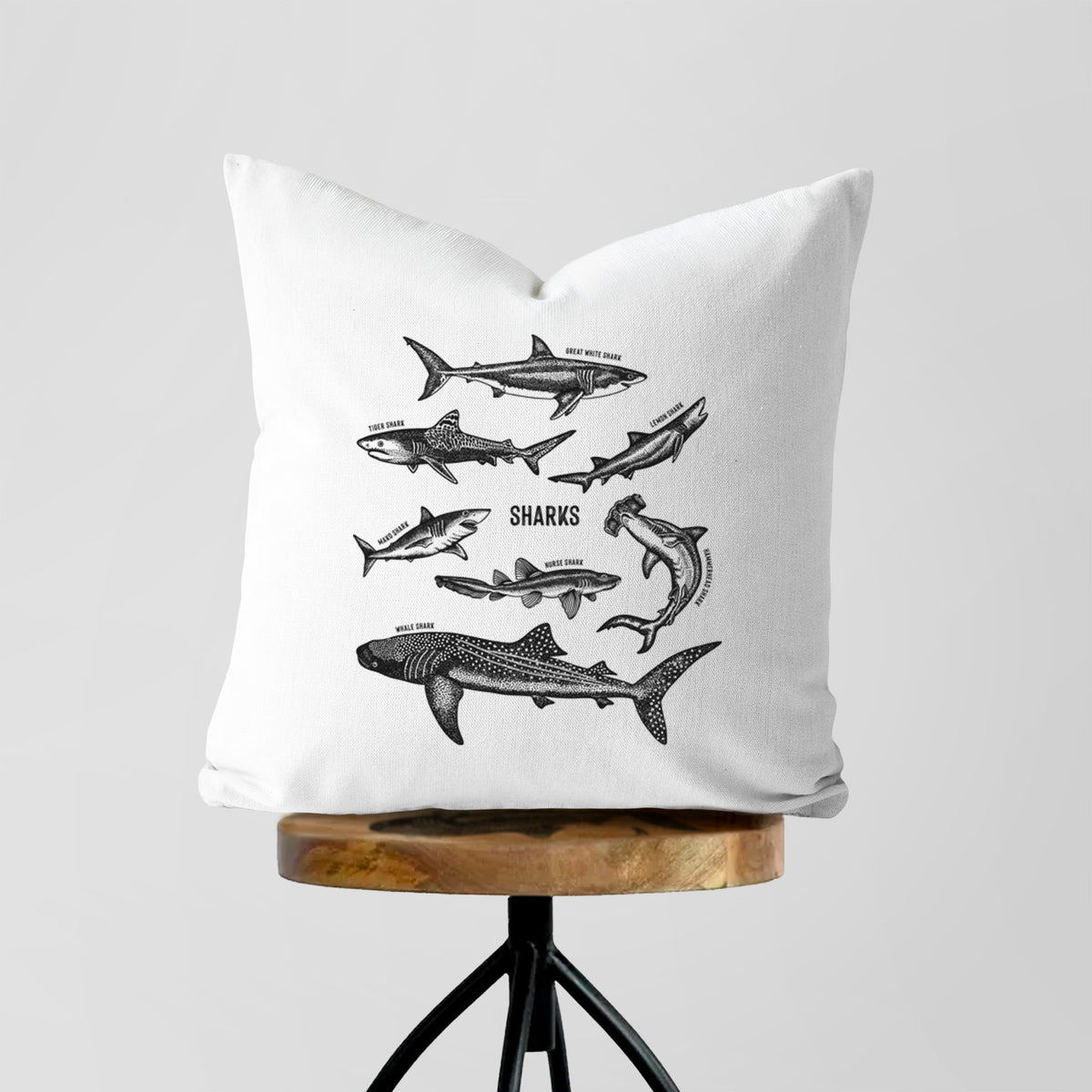 White pillow cover printed with a chart of black ink shark illustrations, including a whale shark, hammerhead and great white, on a wooden stool.