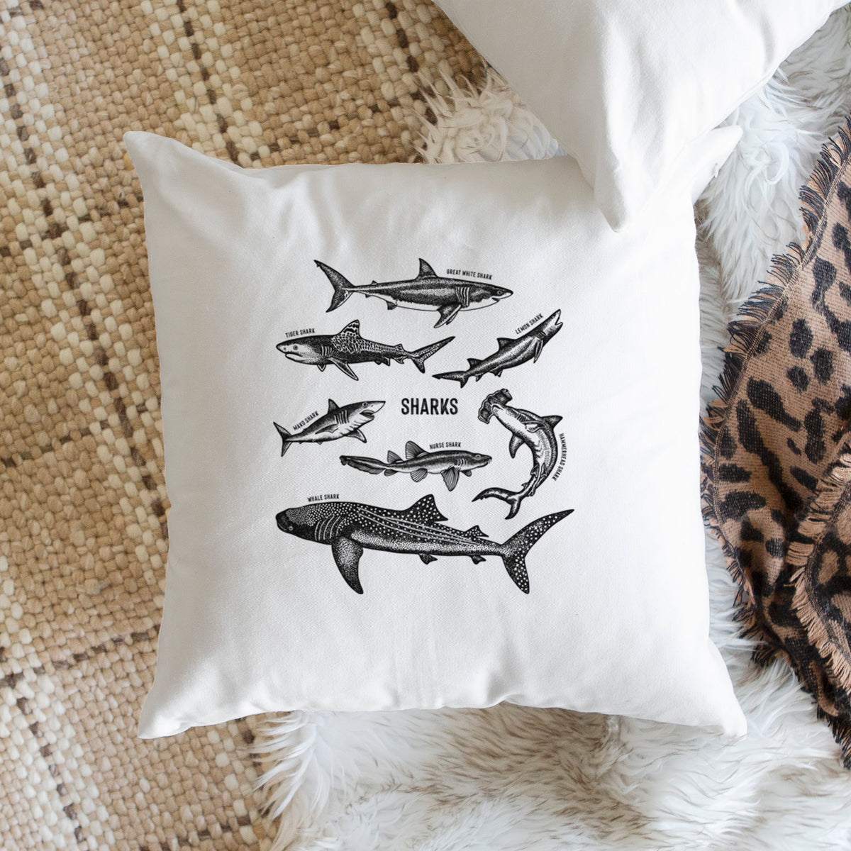 White pillow cover printed with a chart of black ink shark illustrations, including a whale shark, hammerhead and great white, on a woven chair.