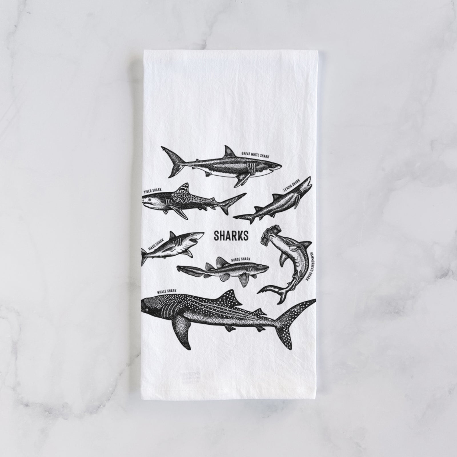 White cotton tea towel printed with a chart of black ink shark illustrations, including a whale shark, hammerhead and great white, on a marble counter beside a round wooden cutting board and greenery.